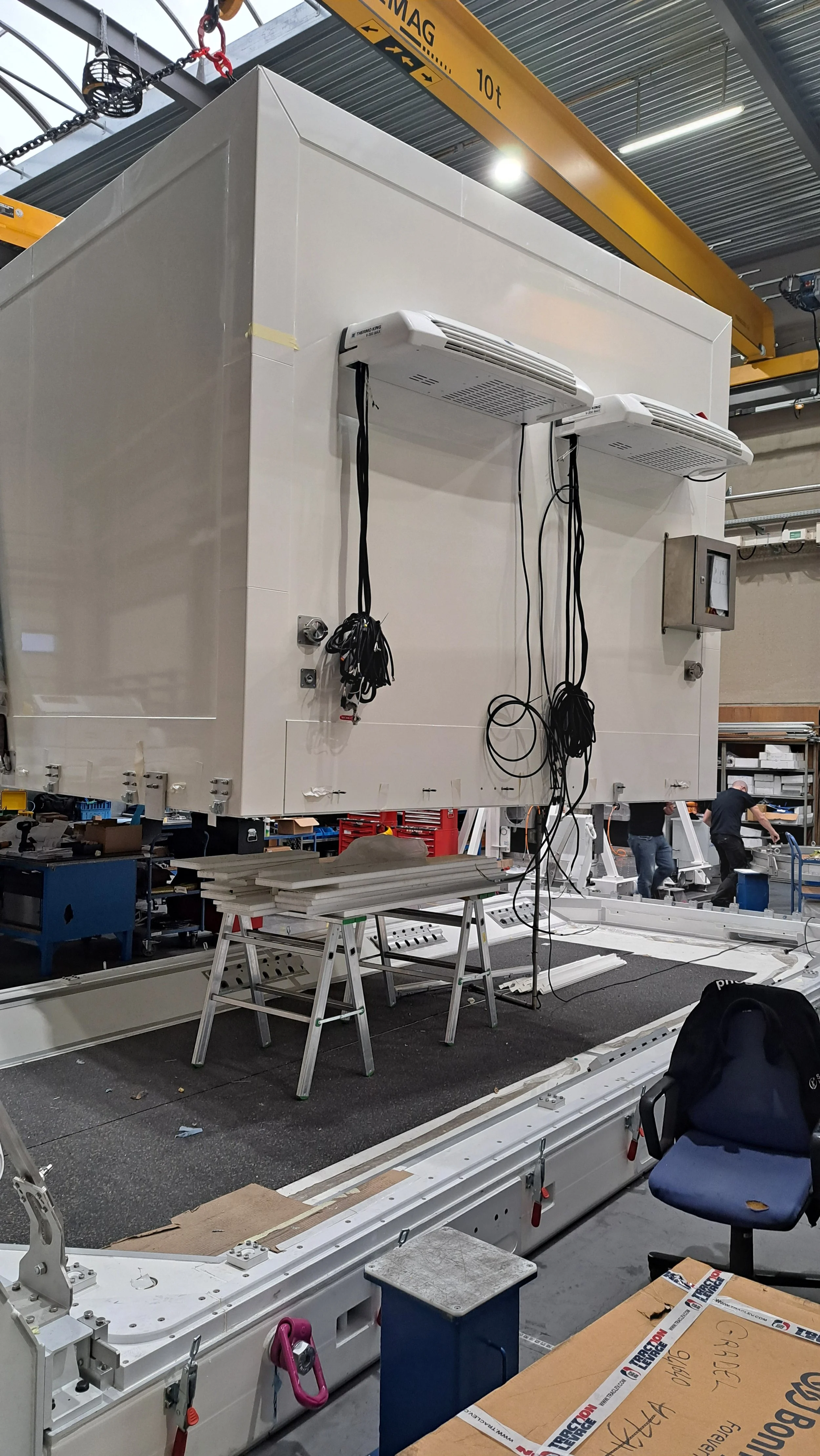 Large white industrial machine or equipment being assembled or installed in a warehouse or factory, with workers and tools in the background.