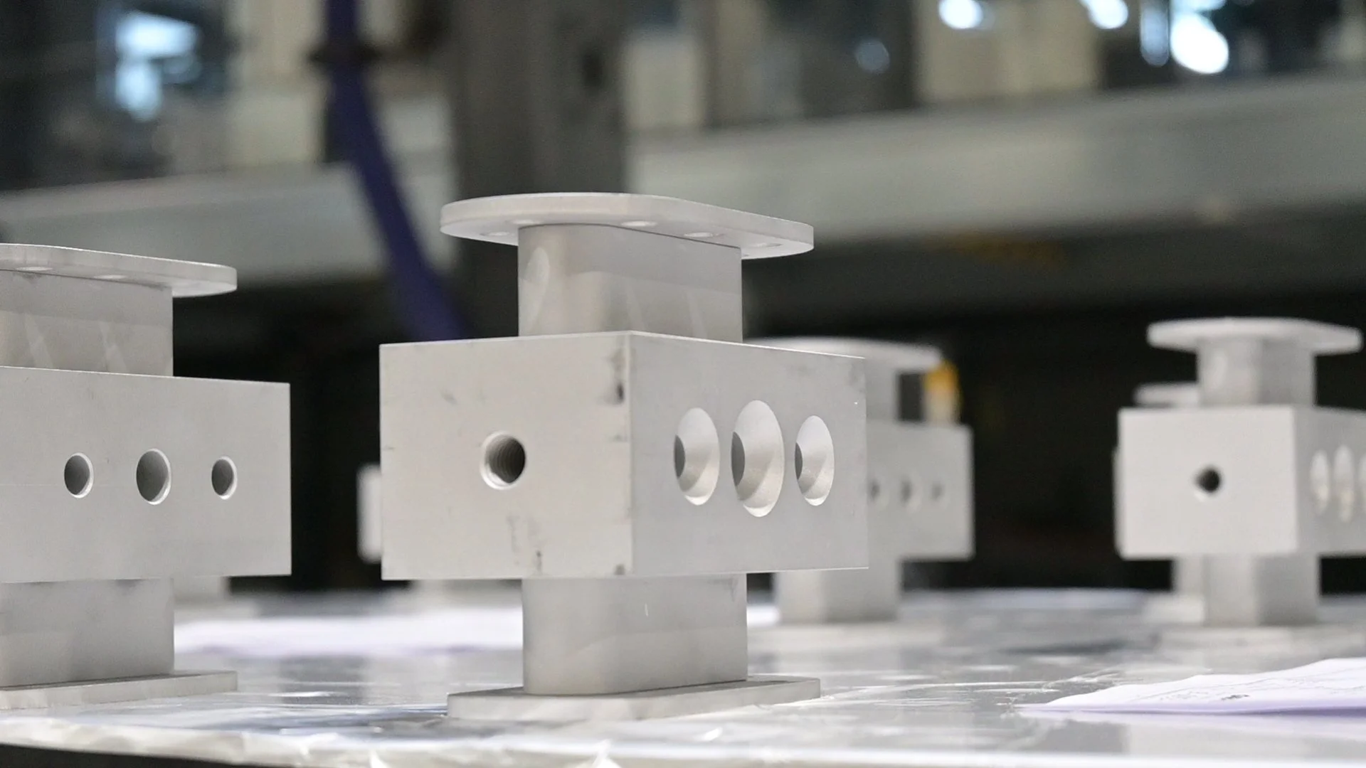 Close-up of metal industrial components with holes and flat top surfaces, arranged on a work surface in a factory setting.
