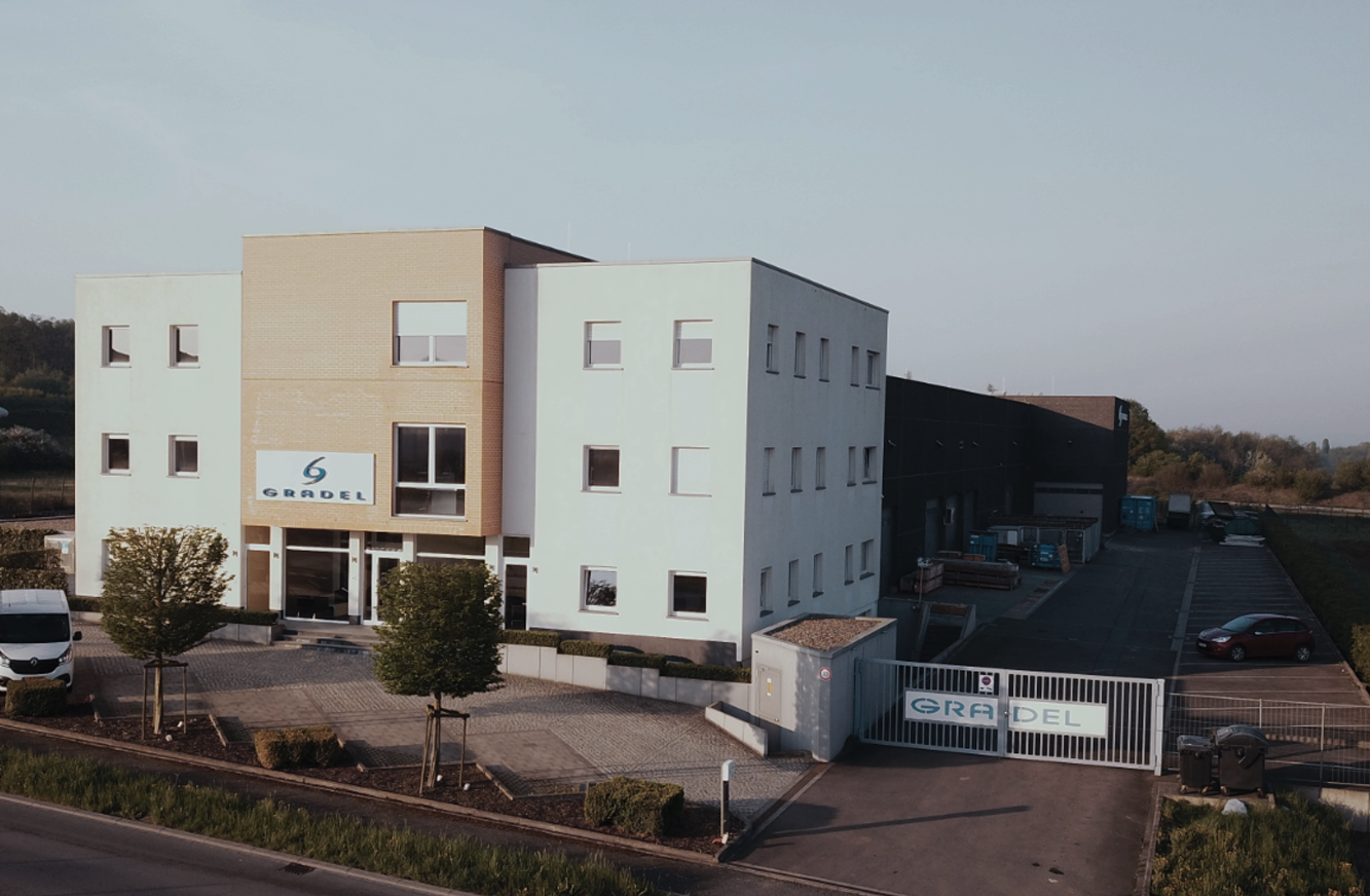 Exterior of a modern industrial building with the logo 'GRADEL' on the front, surrounded by parking lot and trees.