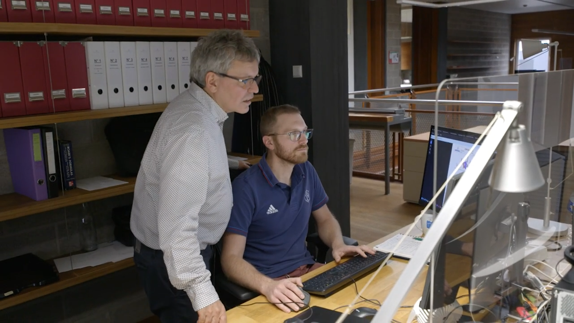 Two men working together at a desk with computer monitors, one standing and one sitting, in an office with wooden floors and shelves of binders in the background.