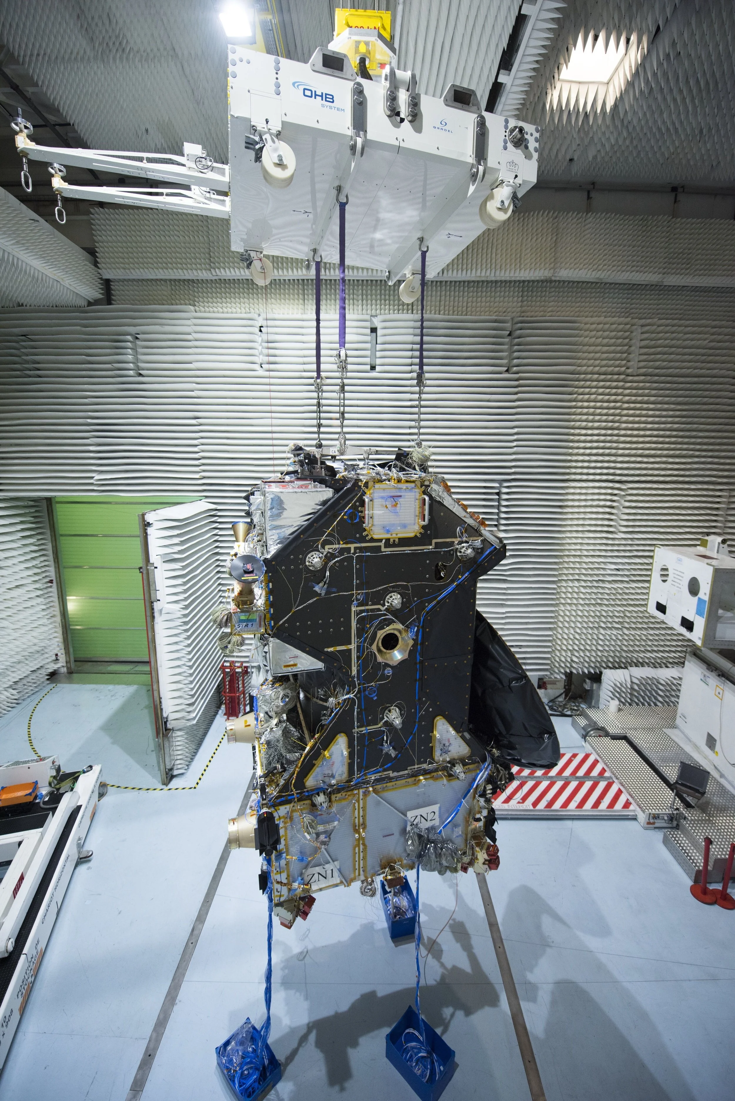 A spacecraft in a clean room, with panels and wires, suspended from the ceiling.