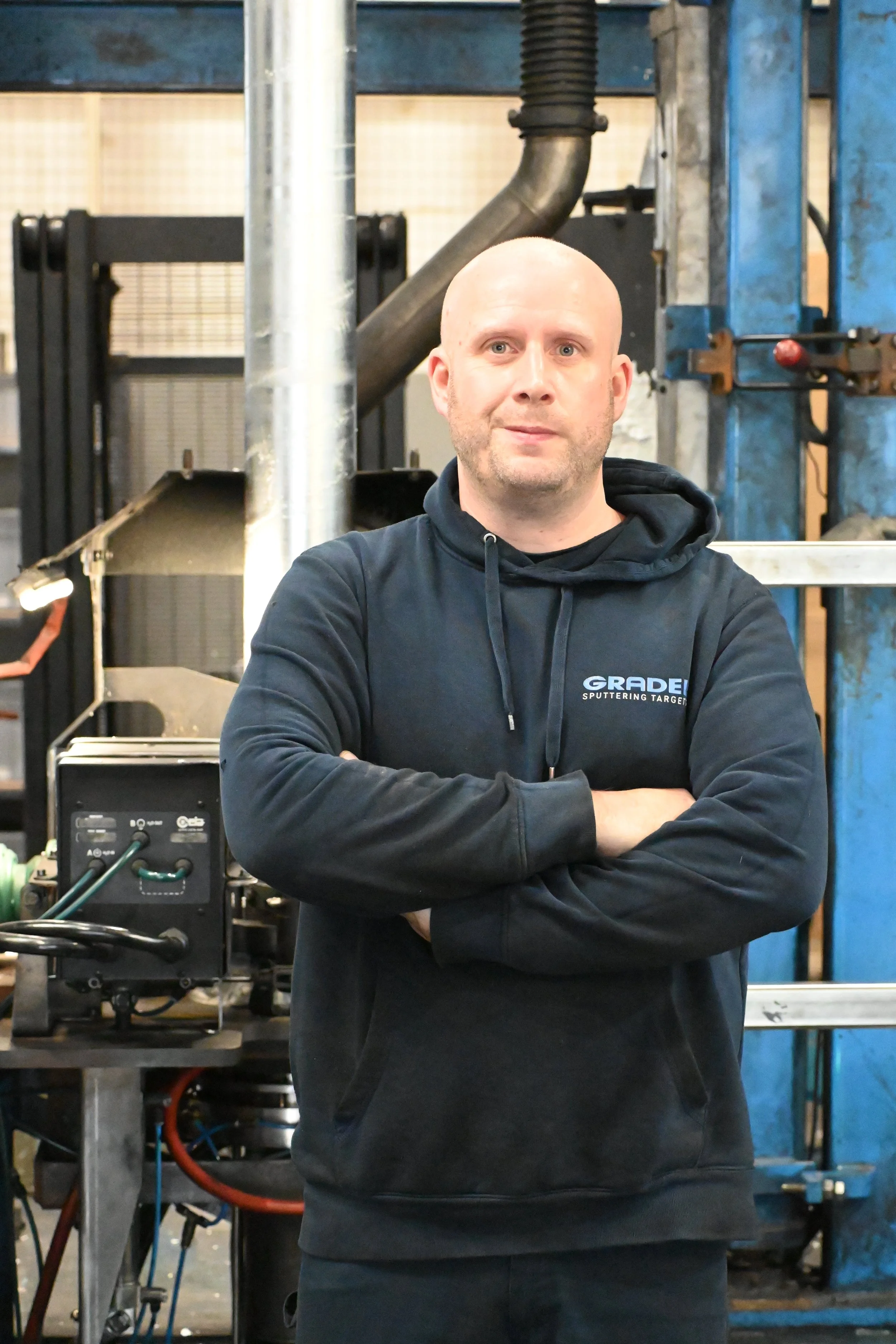 A man with glasses and a beard stands in front of industrial machinery in a factory setting.