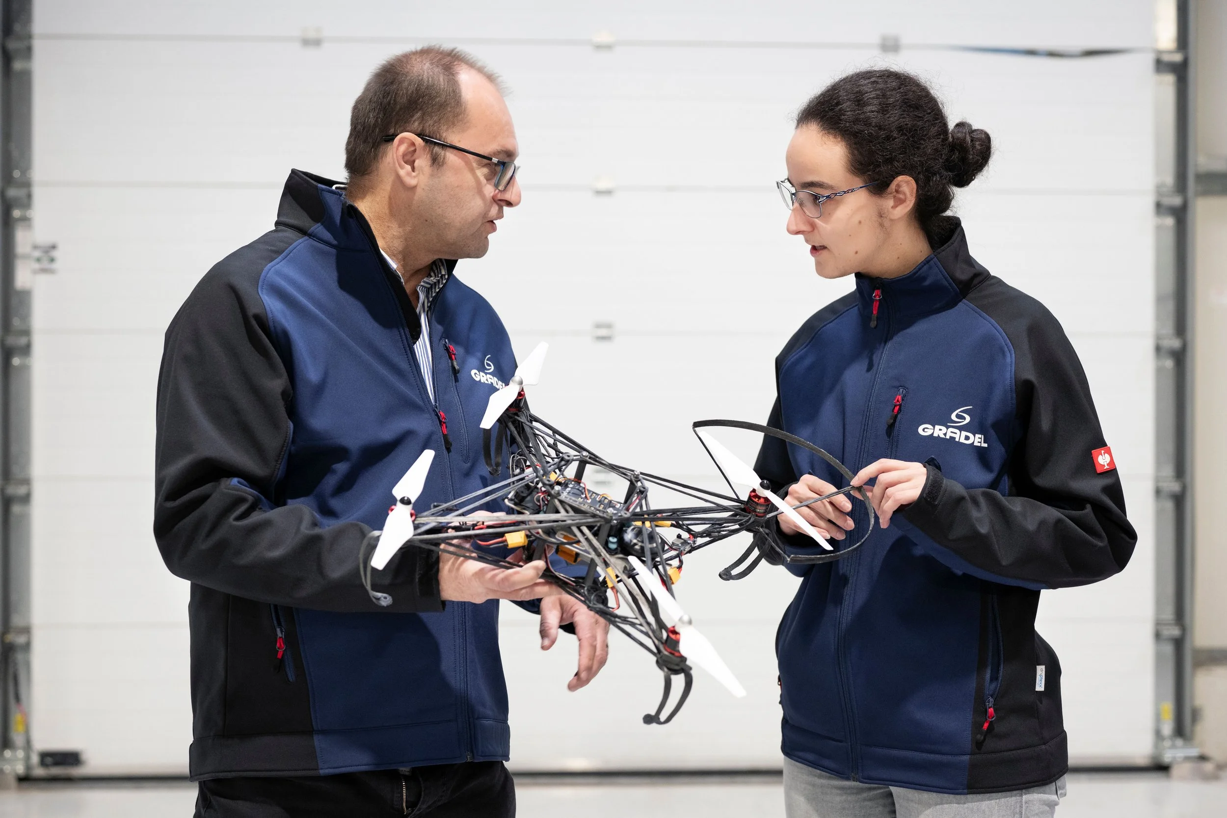 Two people in matching blue and black jackets examining a drone with four white propellers in a workshop or warehouse.
