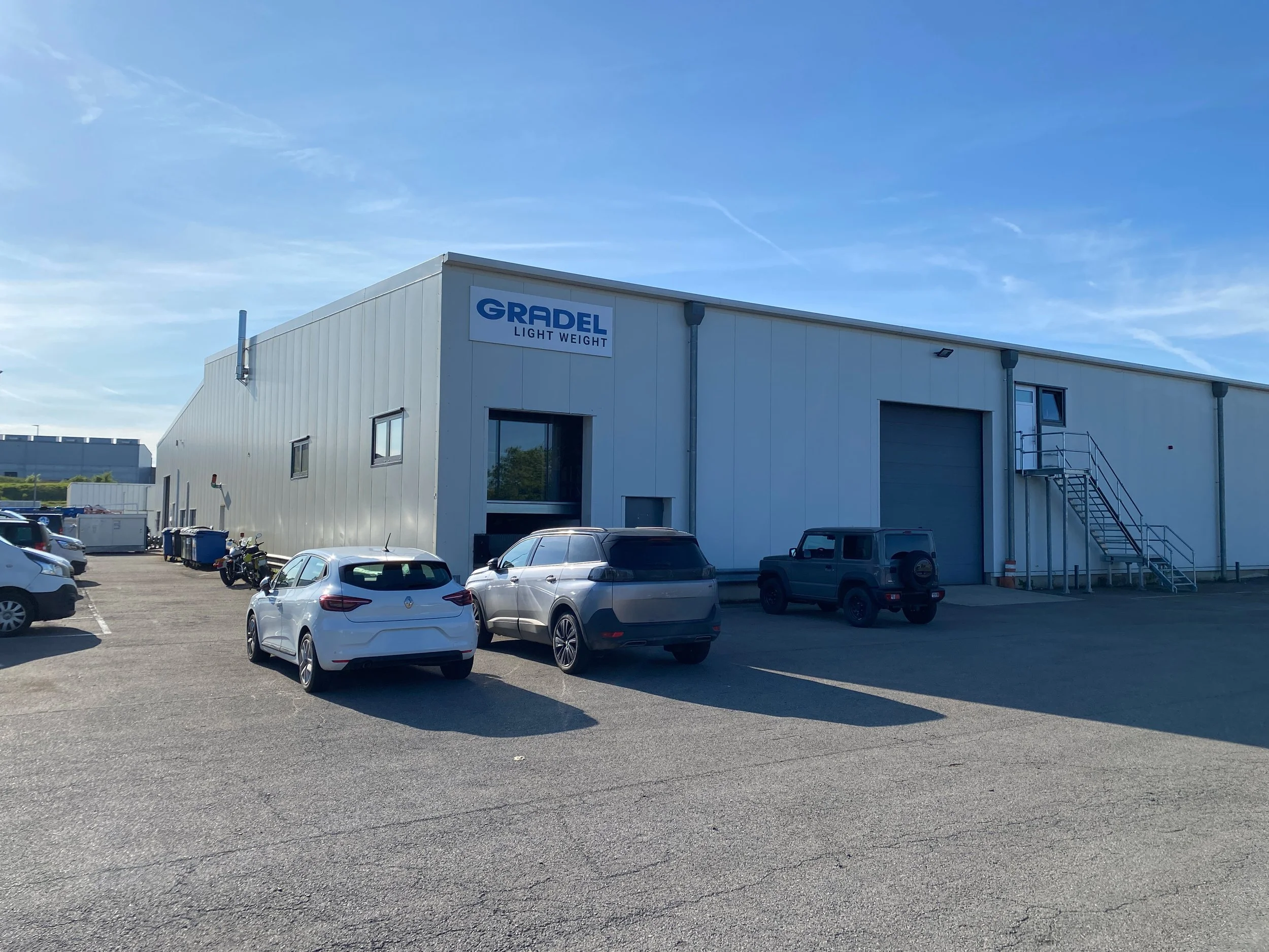 Industrial warehouse building labeled 'GRADEL LIGHT WEIGHT,' with a parking lot in front containing several cars, motorcycles, and trash bins, under a clear blue sky.