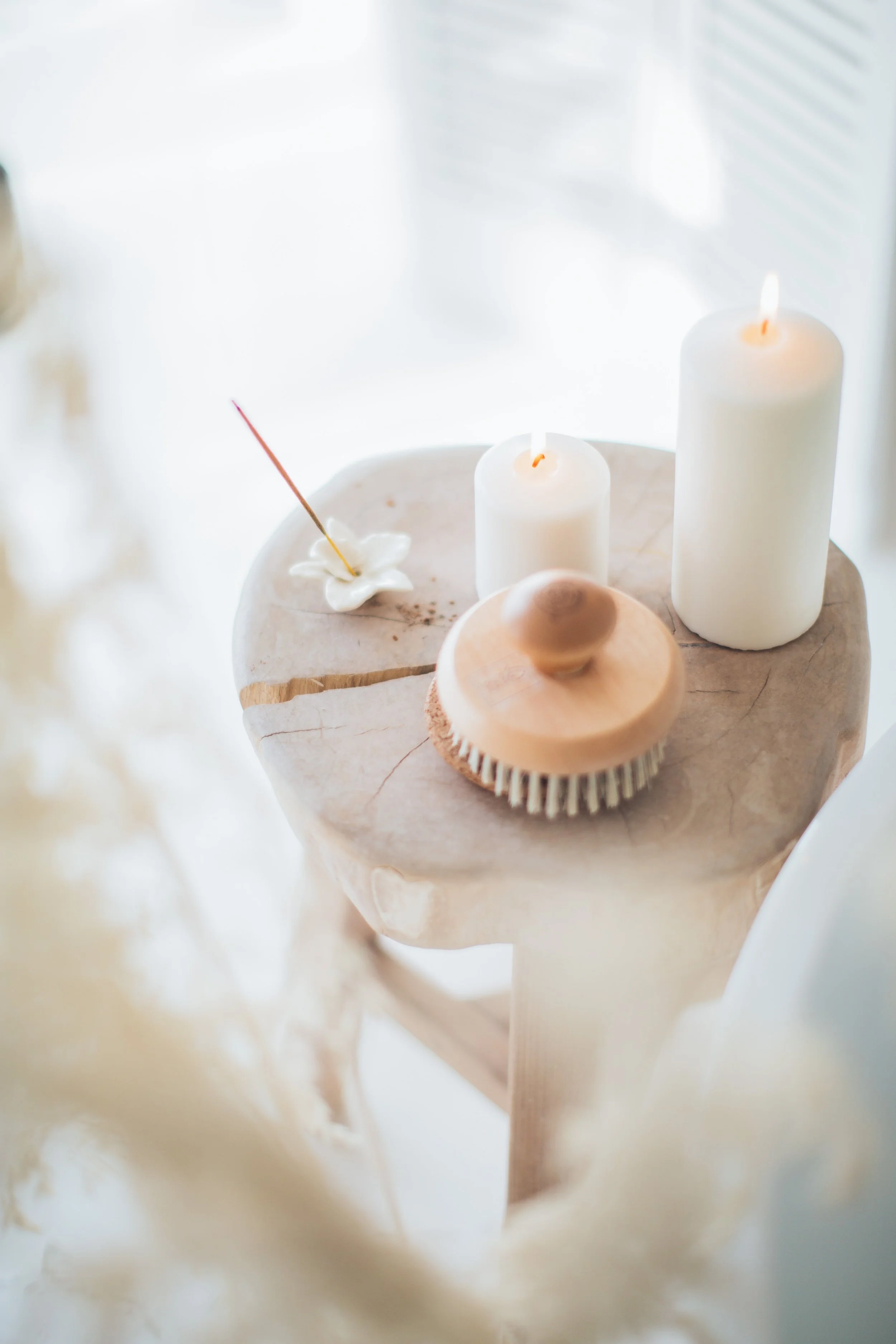 Wooden table with two lit white candles, a wooden body brush, and an incense holder with incense in a bright room.