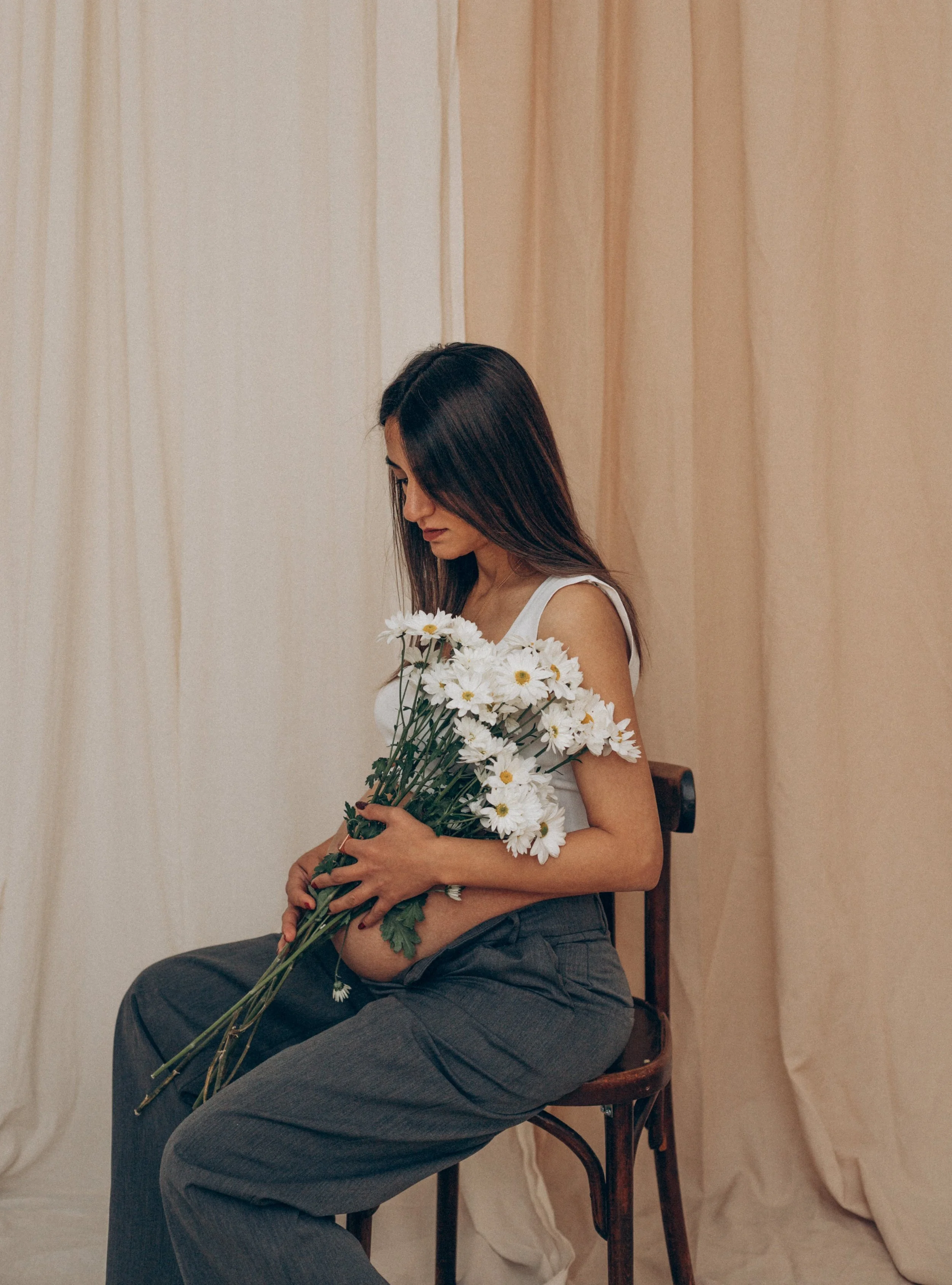 A pregnant woman with long dark hair sitting on a wooden chair, holding a bouquet of white daisies in her lap, against a backdrop of cream-colored curtains.