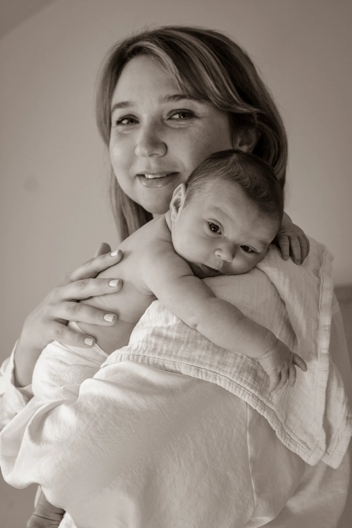 Close-up of a mother with long dark hair holding and nuzzling her newborn baby with reddish hair against her shoulder in a dimly lit setting.