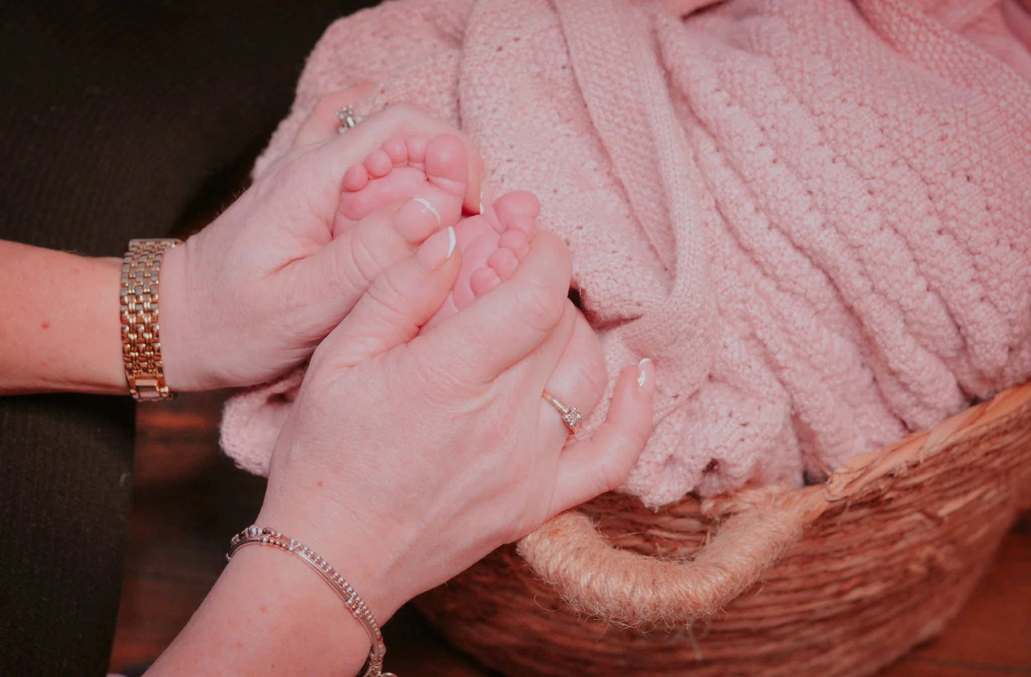 A woman holding a baby's foot, with both wearing rings and the woman also wearing jewelry, in a woven basket lined with pink blanket.