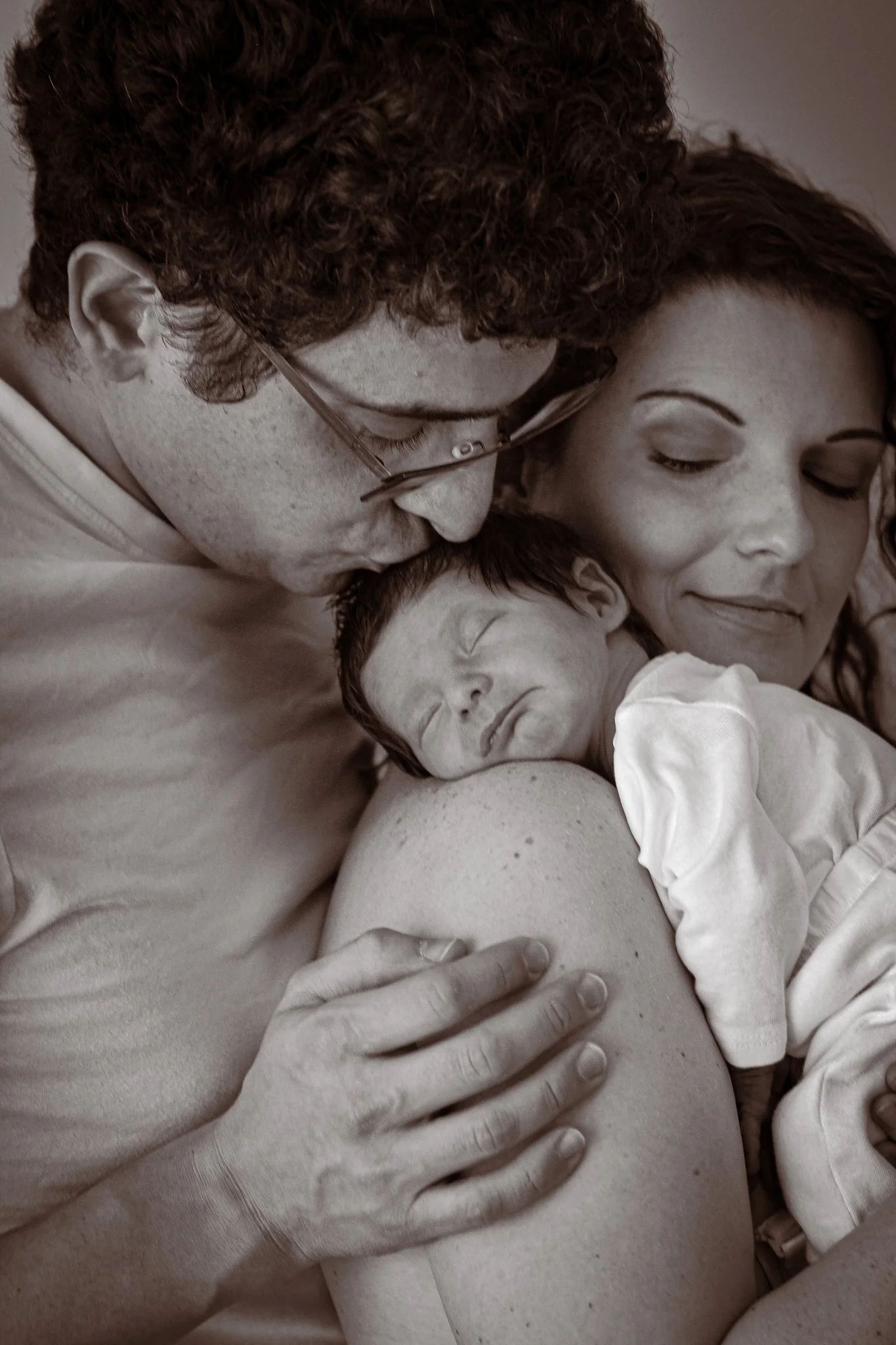 A woman cradles an infant while a man kisses the infant's forehead, all sharing a tender moment.