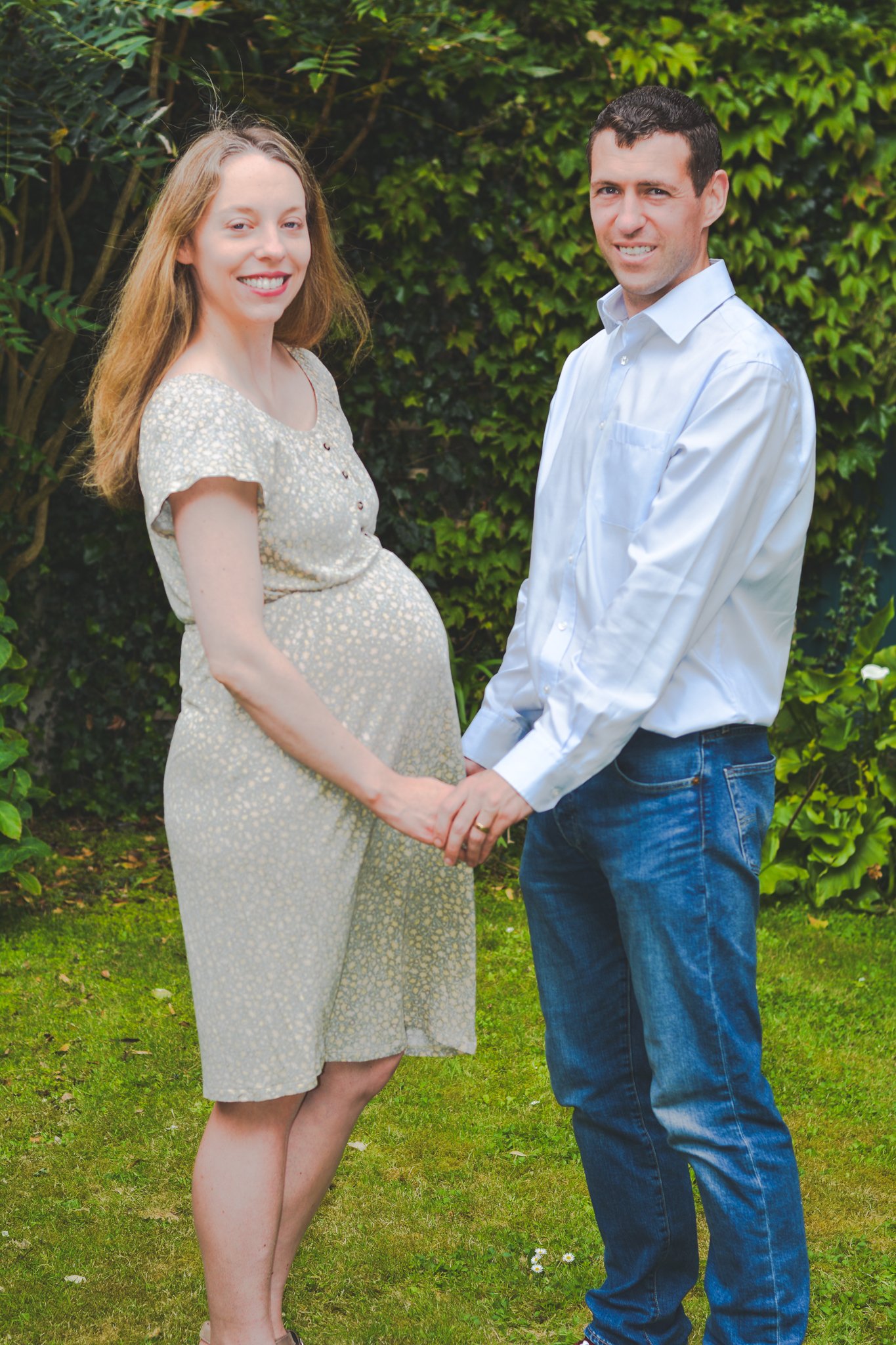 A pregnant woman and a man hold hands and smile outdoors in a garden, with green plants and bushes in the background.