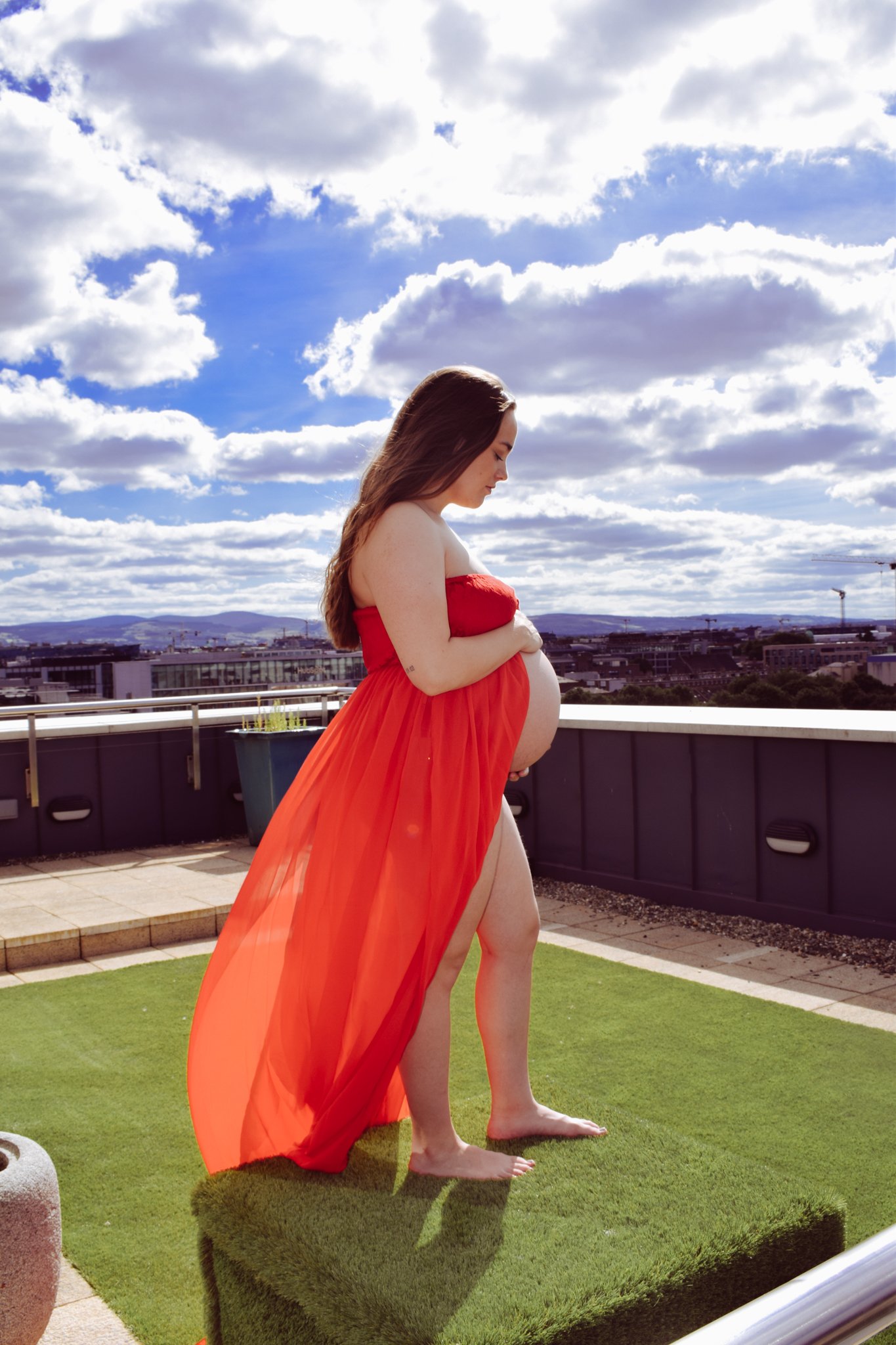A pregnant woman with long brown hair, standing outdoors on a rooftop with a bright sky and clouds. She is wearing a flowing red sheer fabric draped around her, exposing her belly, and appears to be looking down at her stomach in a thoughtful pose.