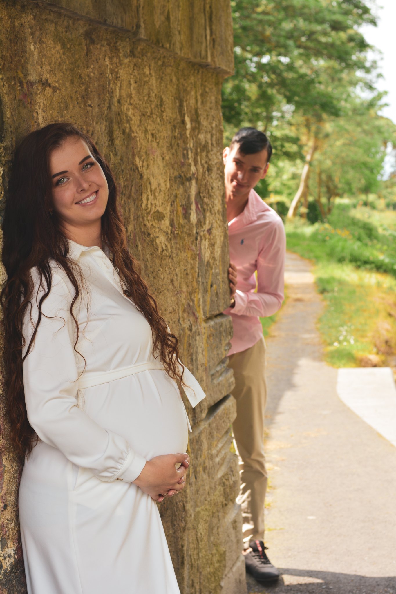A pregnant woman with long brown hair smiling, standing outdoors against a stone wall, and a man peeking from behind the wall, wearing a pink shirt and khaki pants, in a park-like setting with green trees and a pathway.