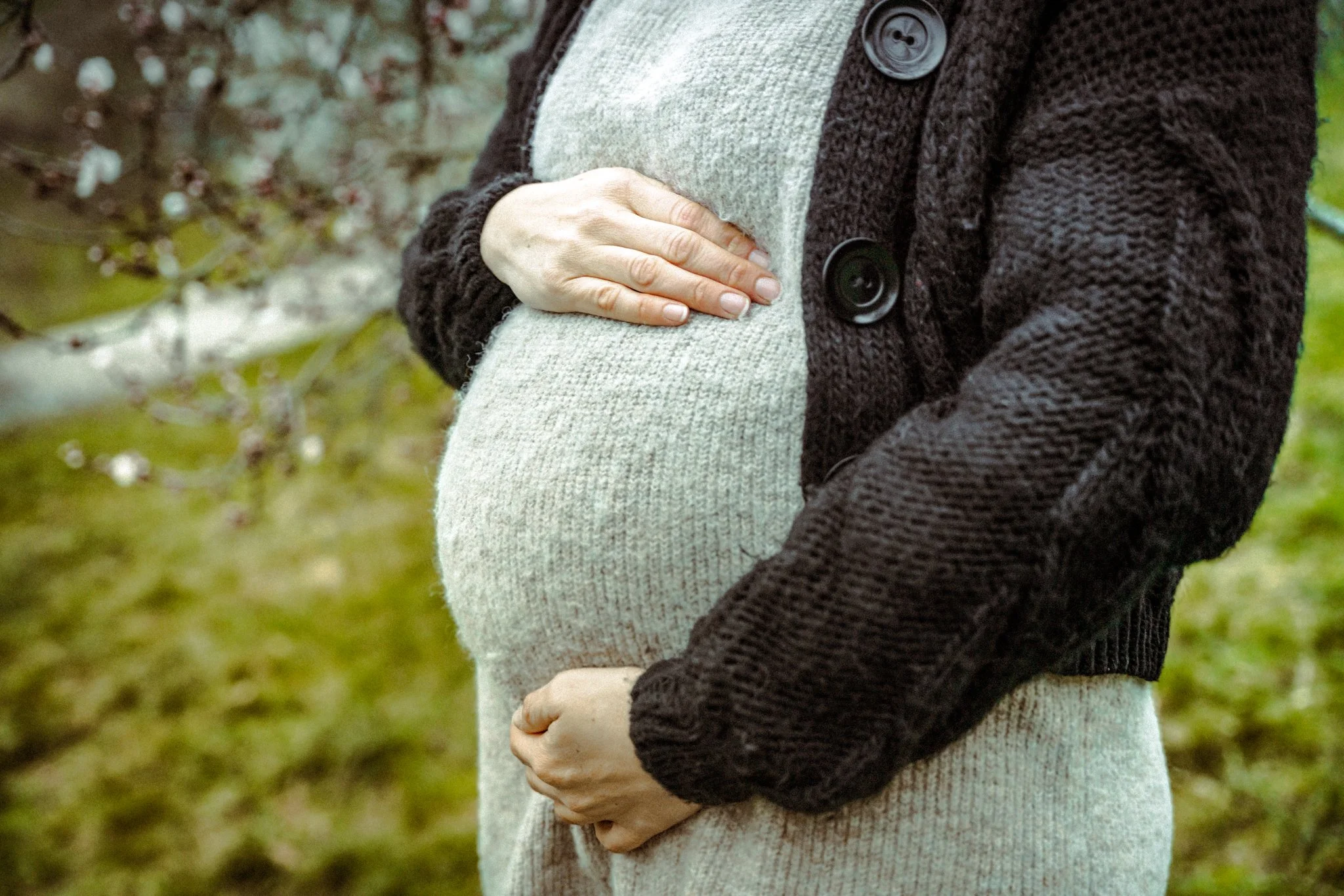 Close-up of a pregnant woman holding her belly with both hands outdoors, wearing a grey sweater and a dark knitted cardigan, with a blurred natural background.