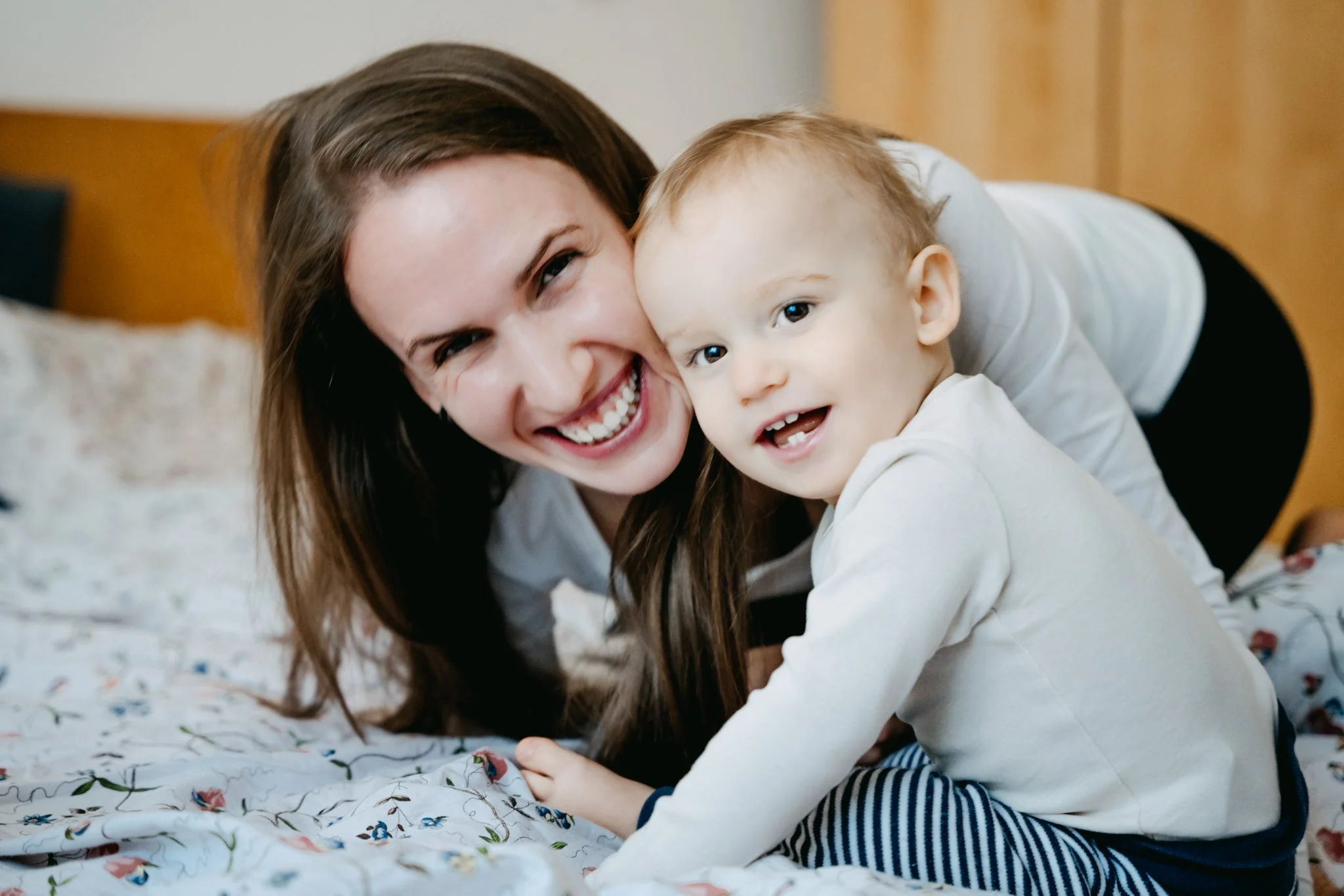 A smiling woman and a young boy play together on a bed with a floral blanket.