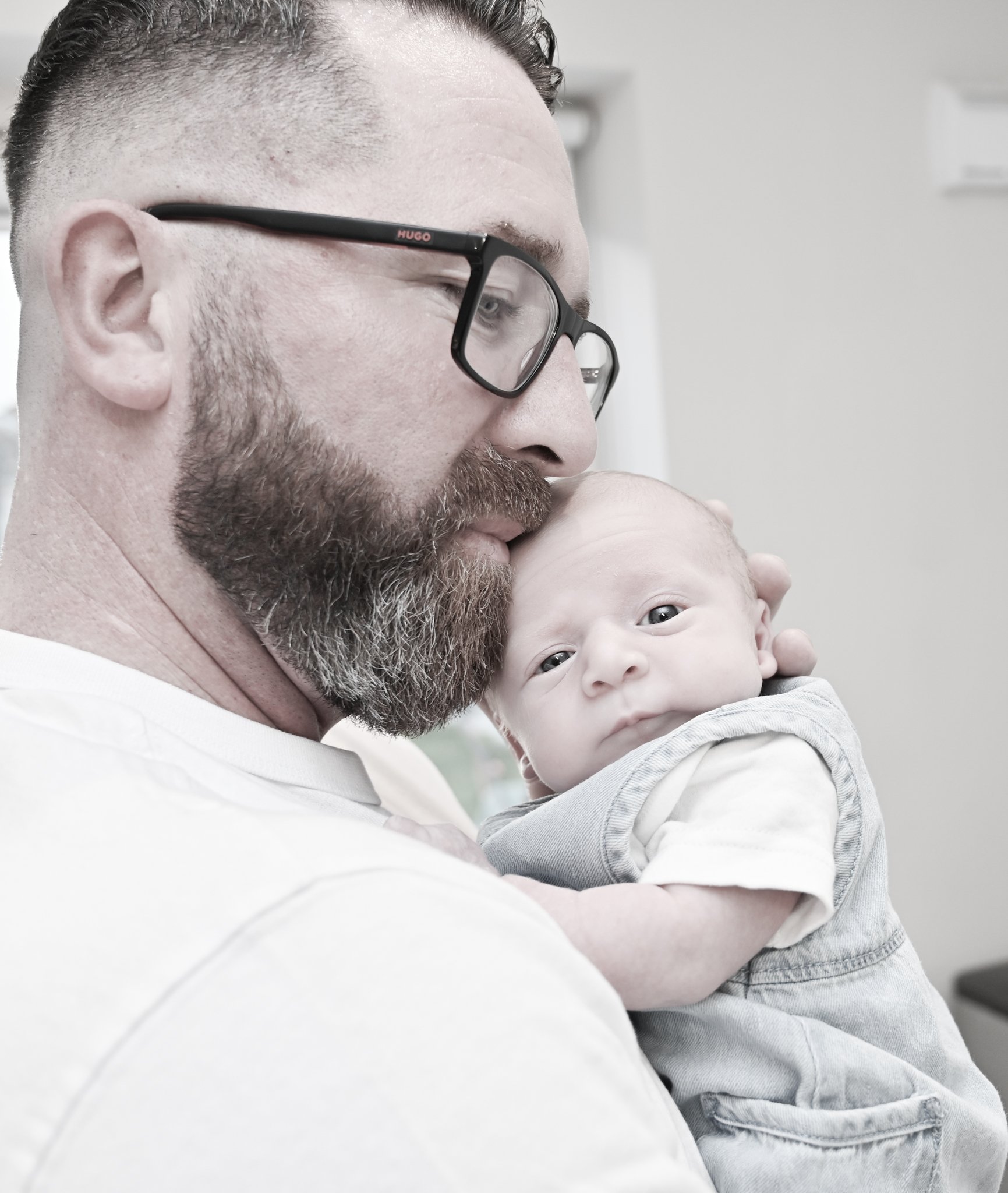 A man with glasses and a beard holding a baby close, with the man gently kissing the baby's forehead.