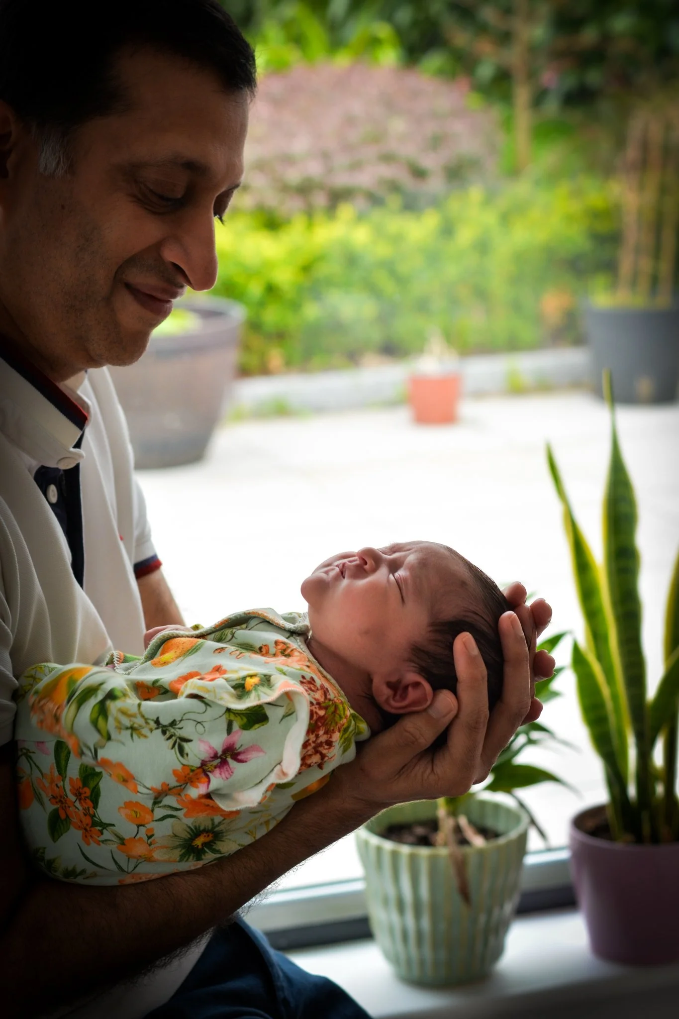 An adult man holding a newborn baby near a window, with potted plants and a garden visible outside.