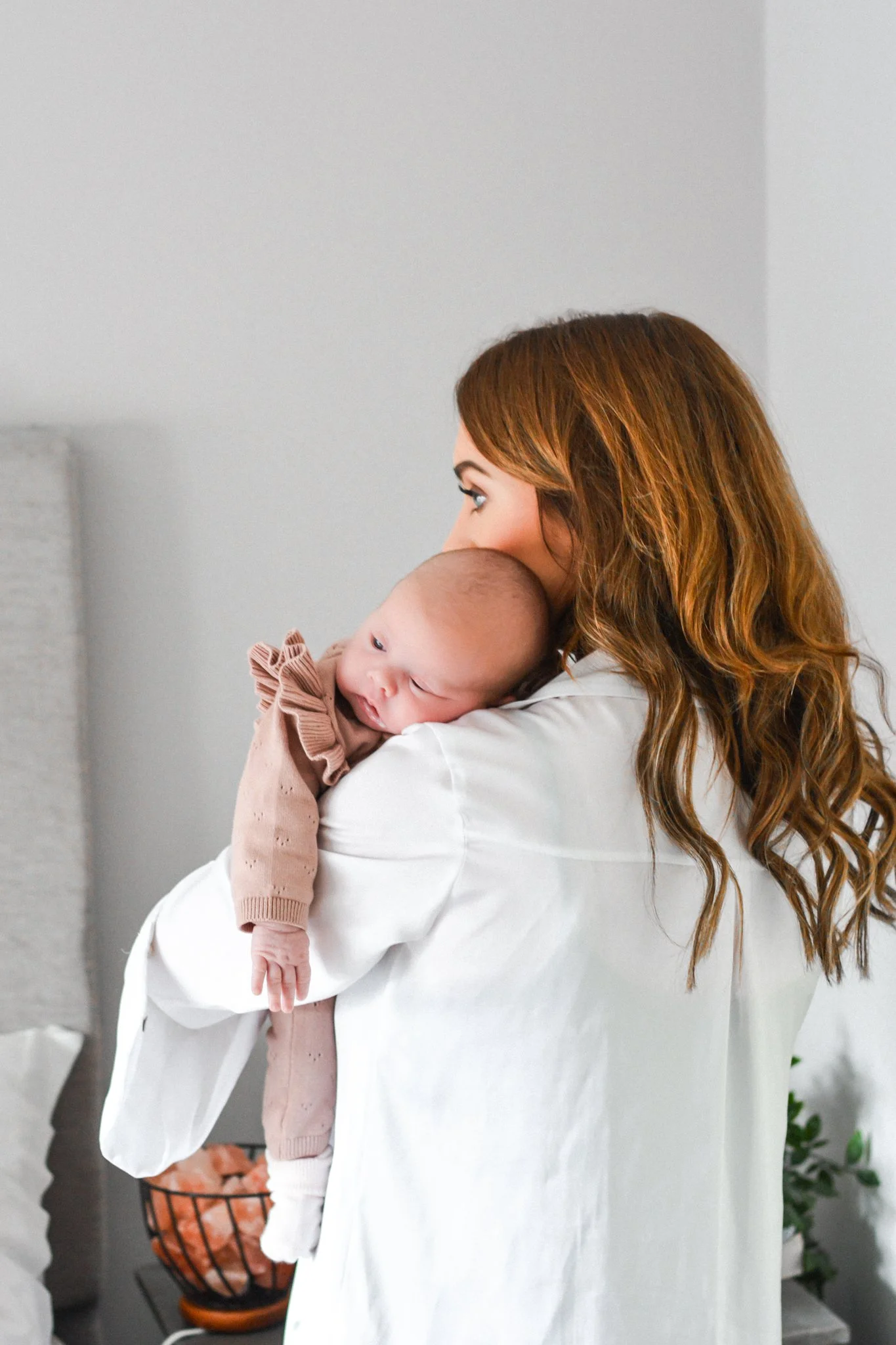 A woman with long, wavy reddish-brown hair holding a baby with light skin and small features close to her shoulder indoors.