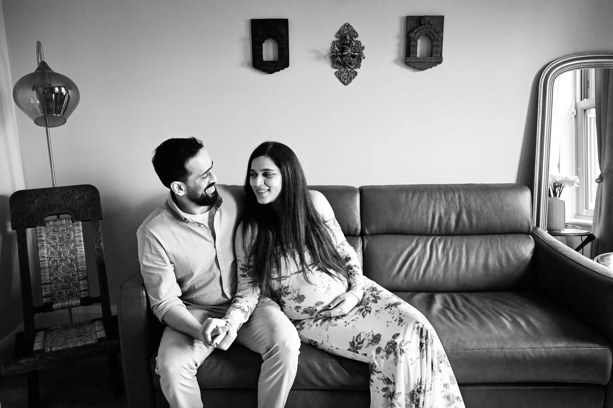 A black and white photo of a man and woman sitting on a sofa, holding hands, smiling, and looking at each other in a cozy living room.