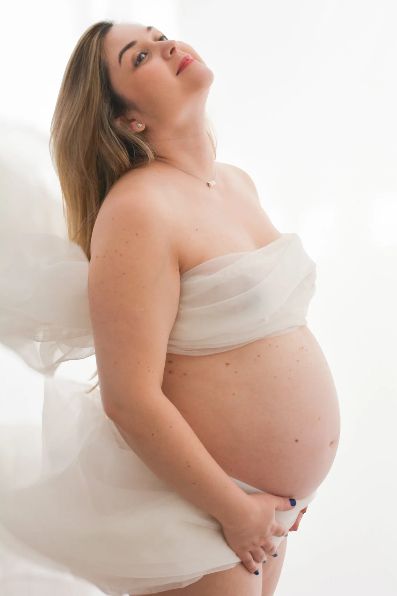 Pregnant woman with long hair, standing against a white background, partially draped in a sheer white fabric, holding her belly with one hand.