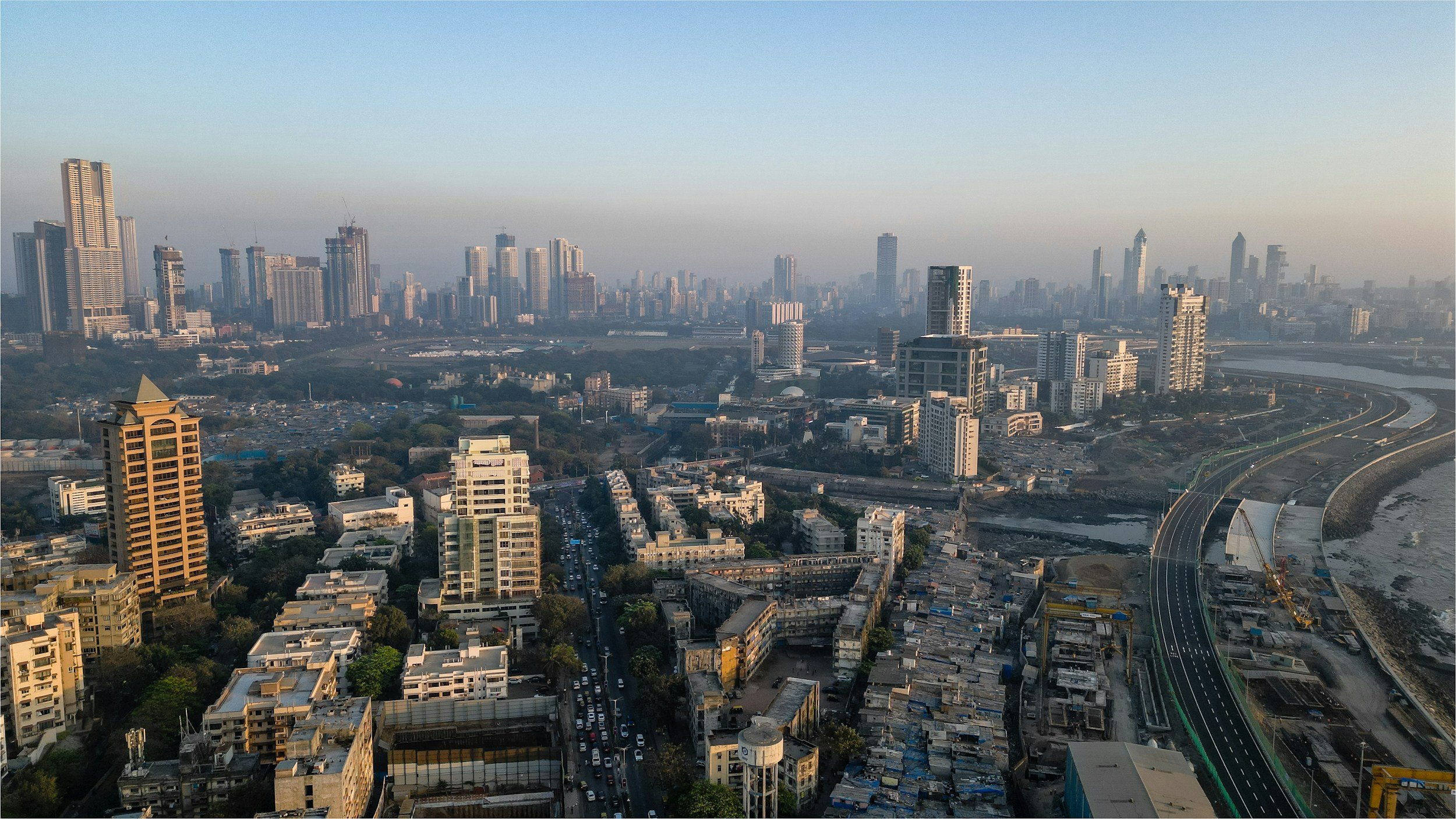 A cityscape with tall buildings, a highway under construction, and a river on the right. The scene is hazy, with a clear sky and early morning or late afternoon lighting.