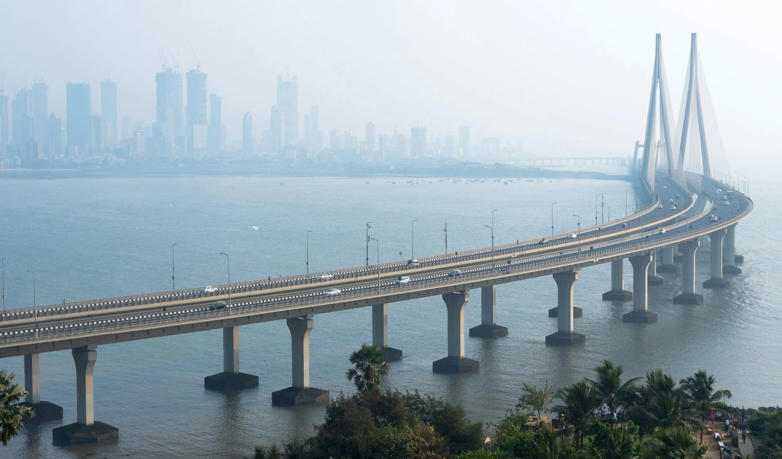 A large bridge spanning over water with a city skyline in the background, featuring tall skyscrapers under a hazy sky.