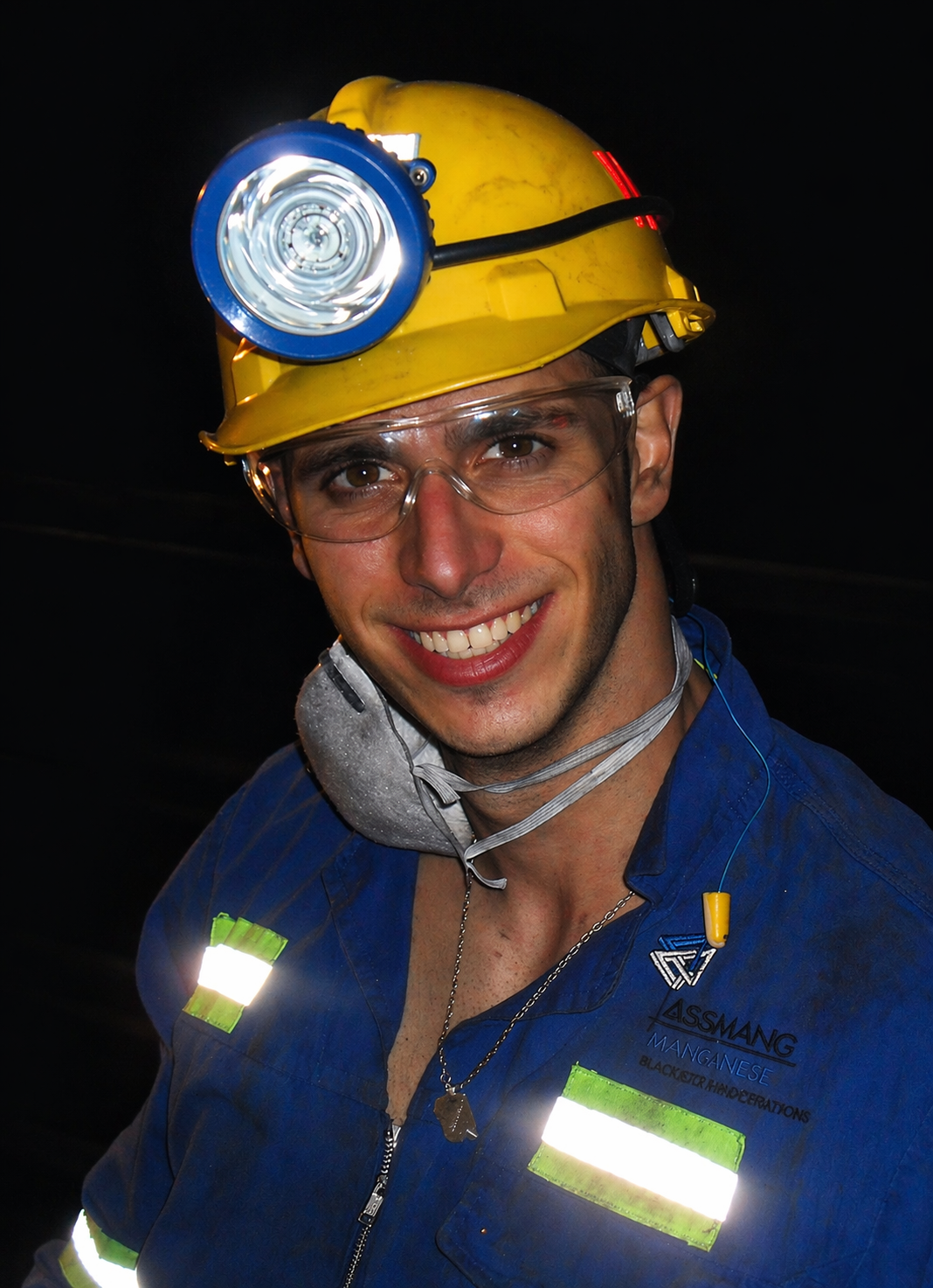 Nicholas Vafeas smiling, wearing a yellow hard hat with a headlamp, safety glasses, and a blue uniform with reflective strips, standing in an underground mine.