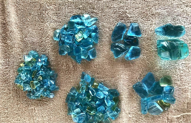 Various sizes of broken blue-green glass pieces arranged on a textured beige surface.