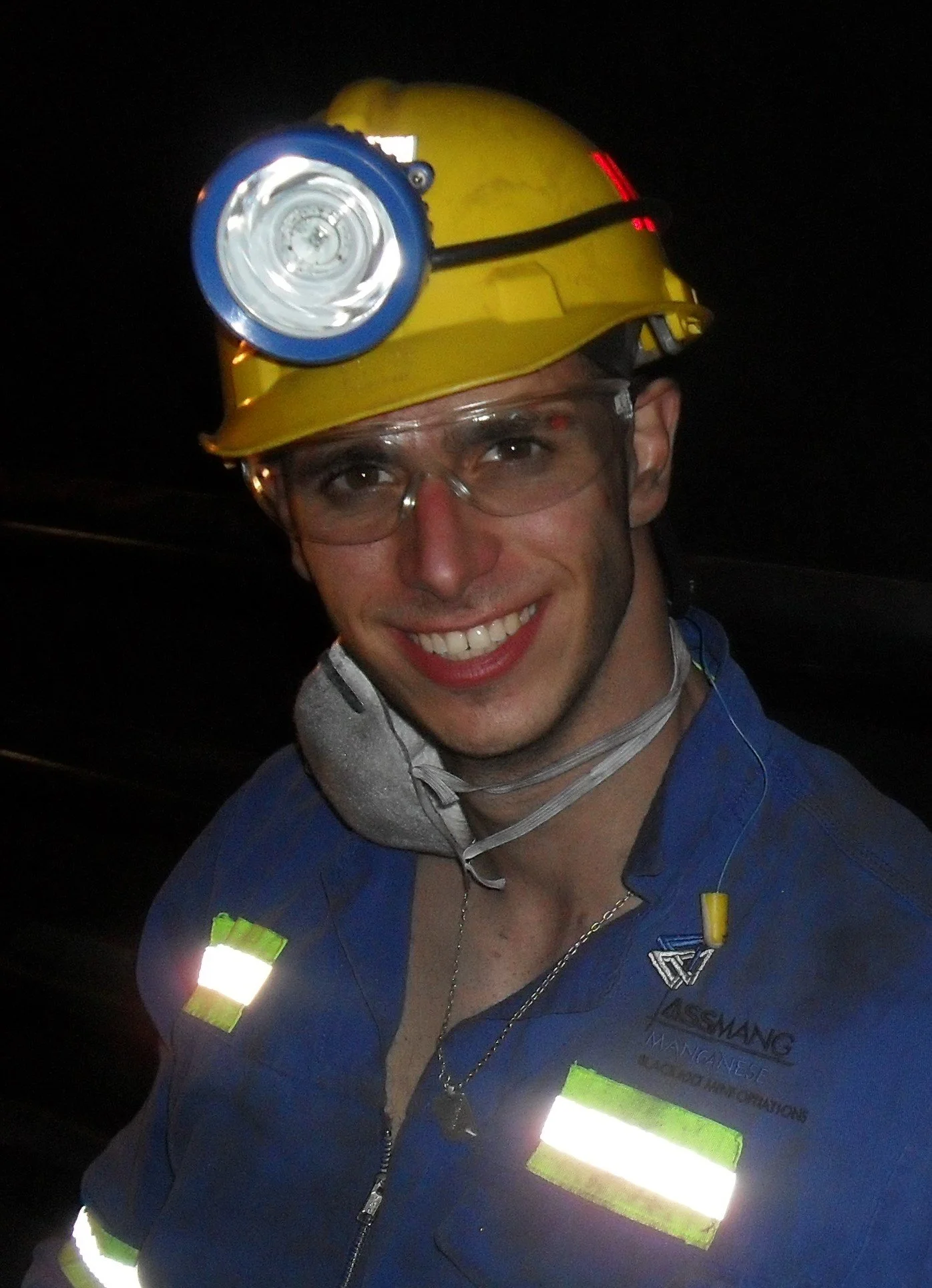 A smiling man wearing a yellow safety helmet with a mining headlamp, safety glasses, and a blue work uniform with reflective patches.