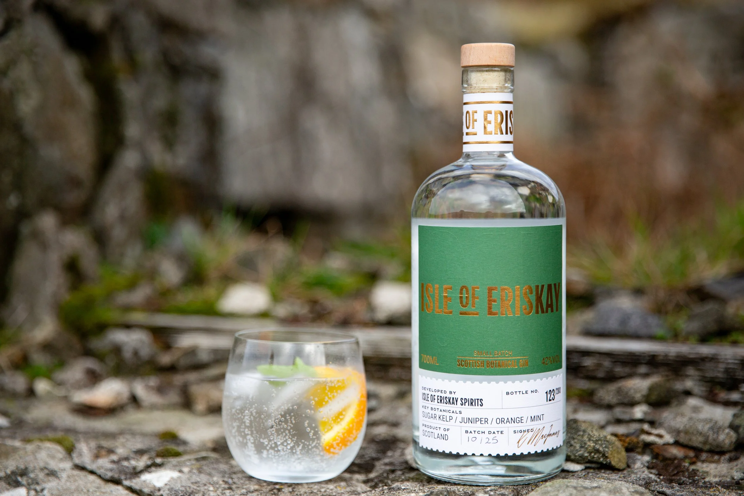 A clear bottle of Isle of Eriskay Scottish botanical gin with a cork cap, placed on a rocky surface outdoors, alongside a glass of gin and tonic with lemon and mint slices.