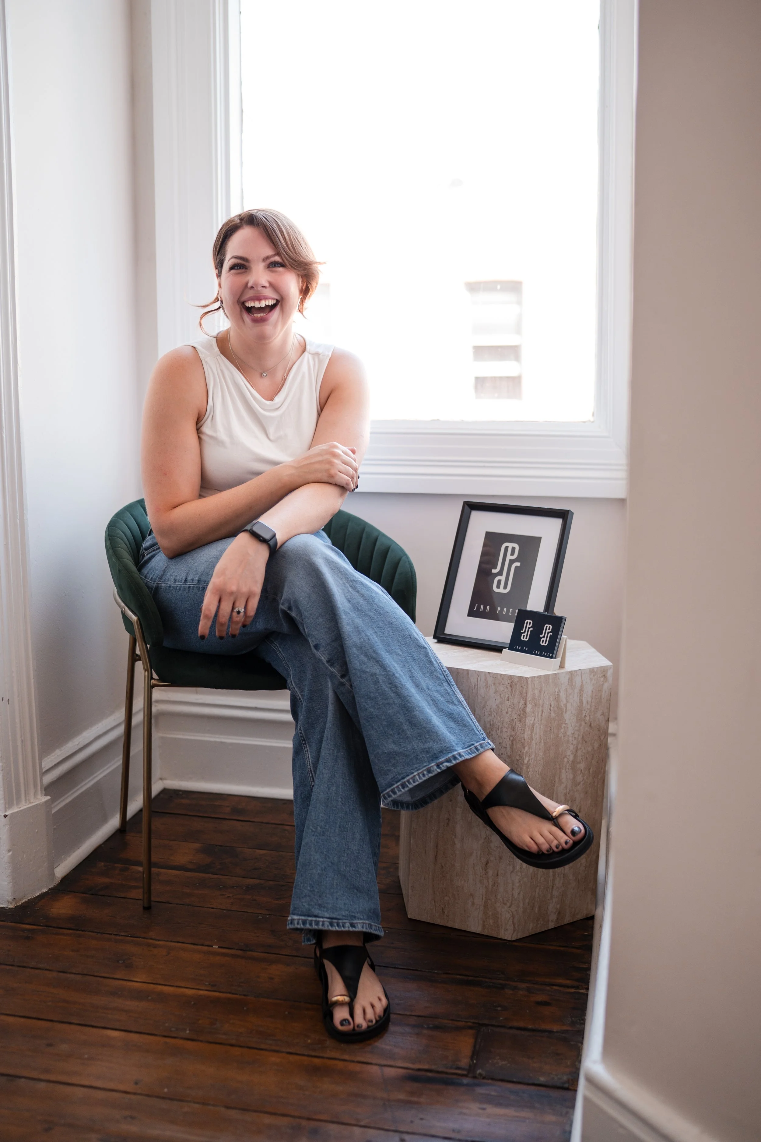 A woman sitting on a green chair, laughing, with a framed picture and a small piece of art on a side table near a window.