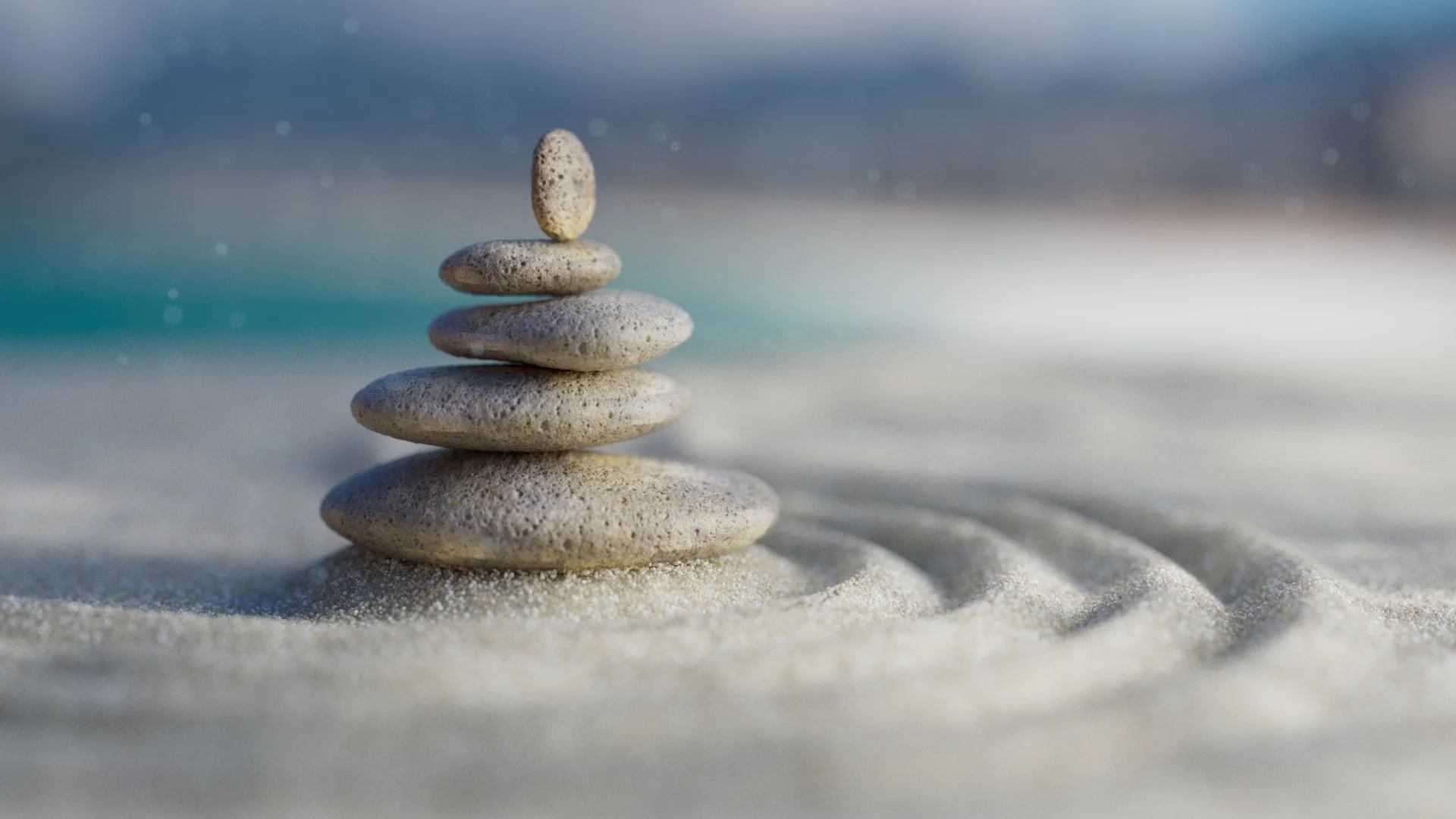 Five stacked smooth stones on sandy beach with water and cloudy sky in the background.