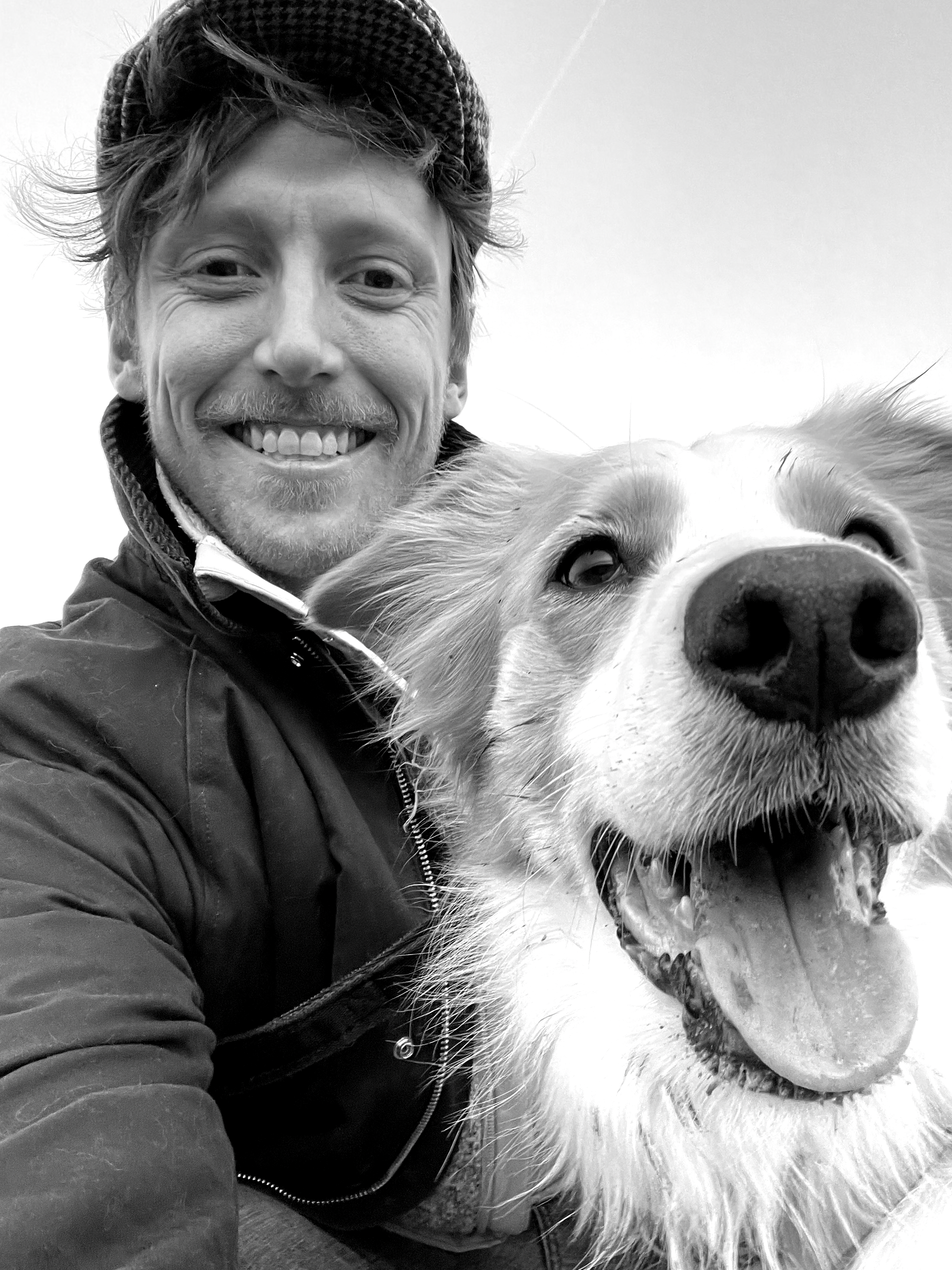 A man smiling for a selfie with a happy dog with its mouth open. The man is wearing a jacket and a knit hat.
