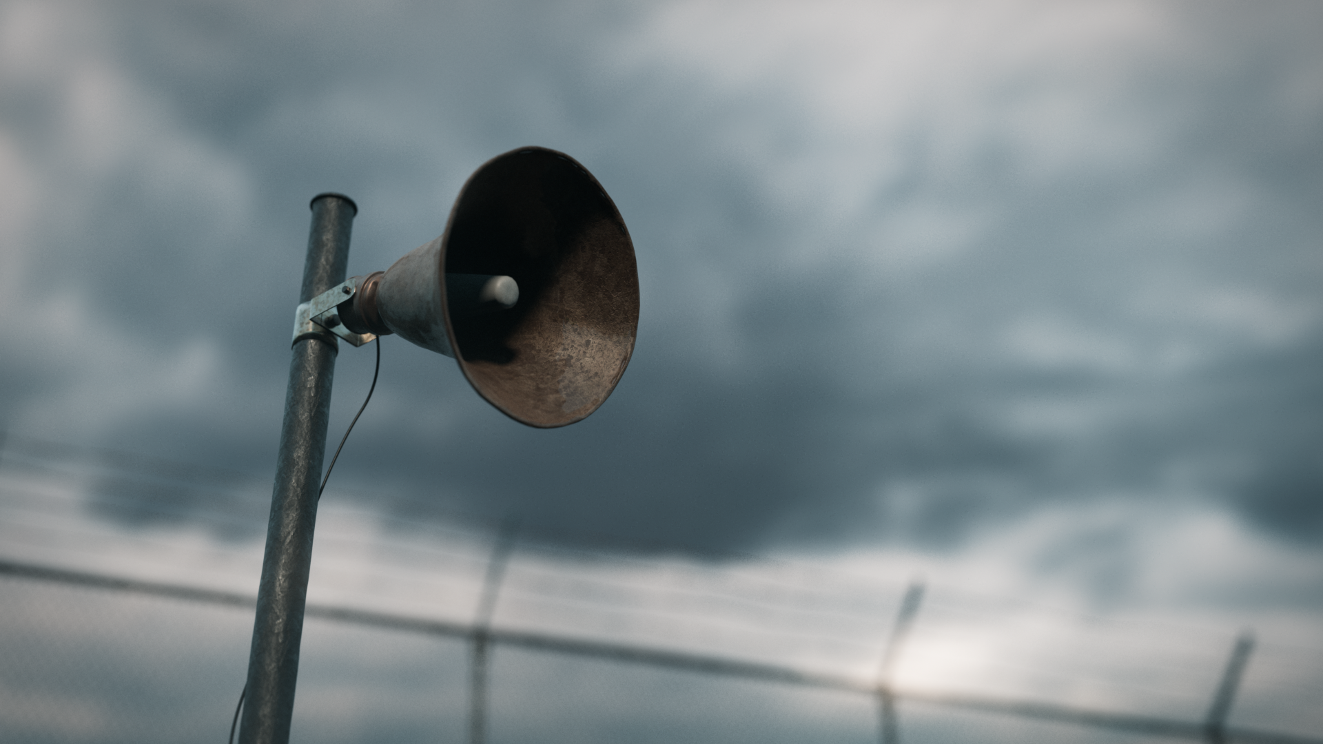 An old, rusty outdoor loudspeaker mounted on a metal pole against a cloudy, overcast sky.