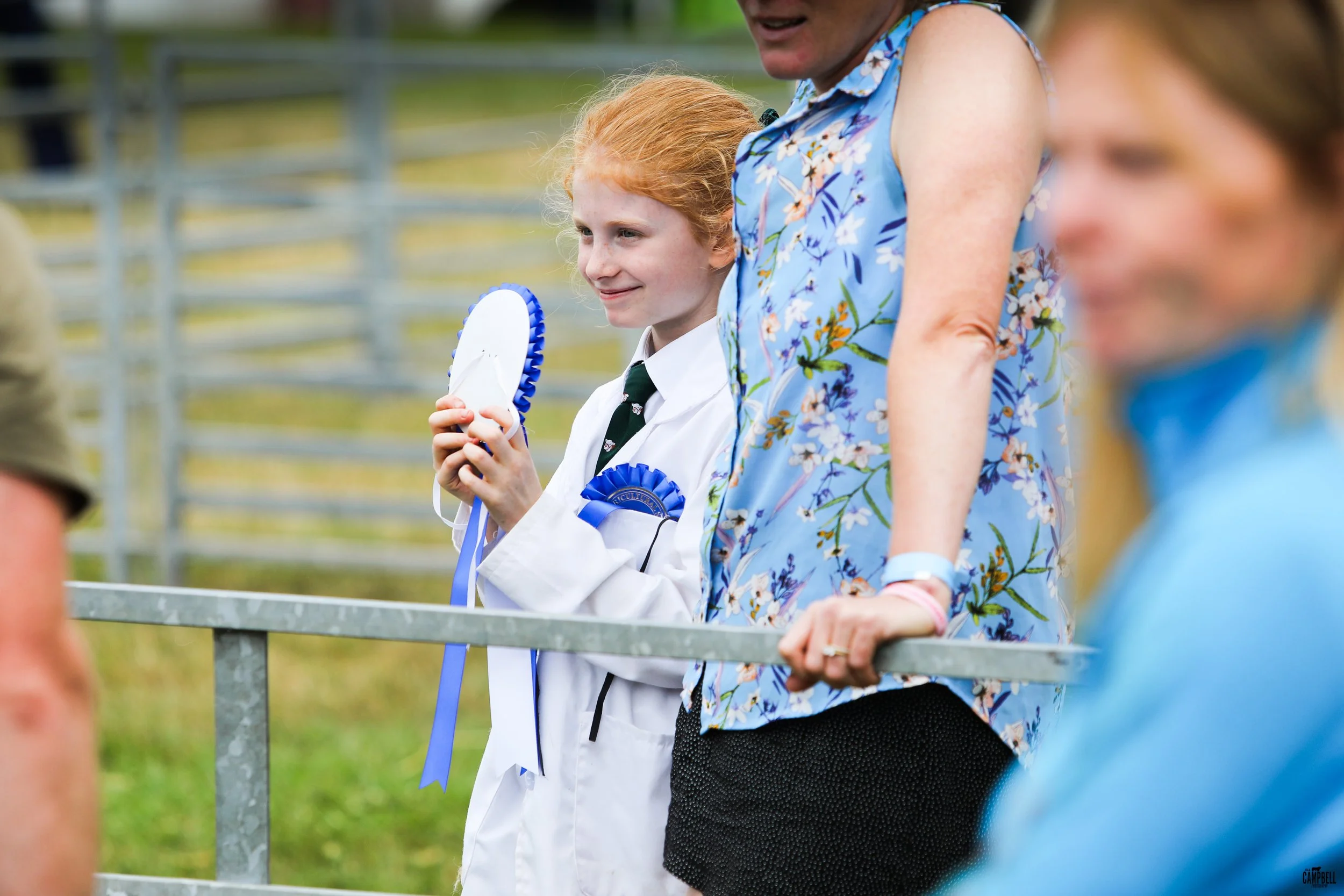 A young girl with red hair holding a blue and white ribbon at an outdoor event, with people and a metal fence in the background.