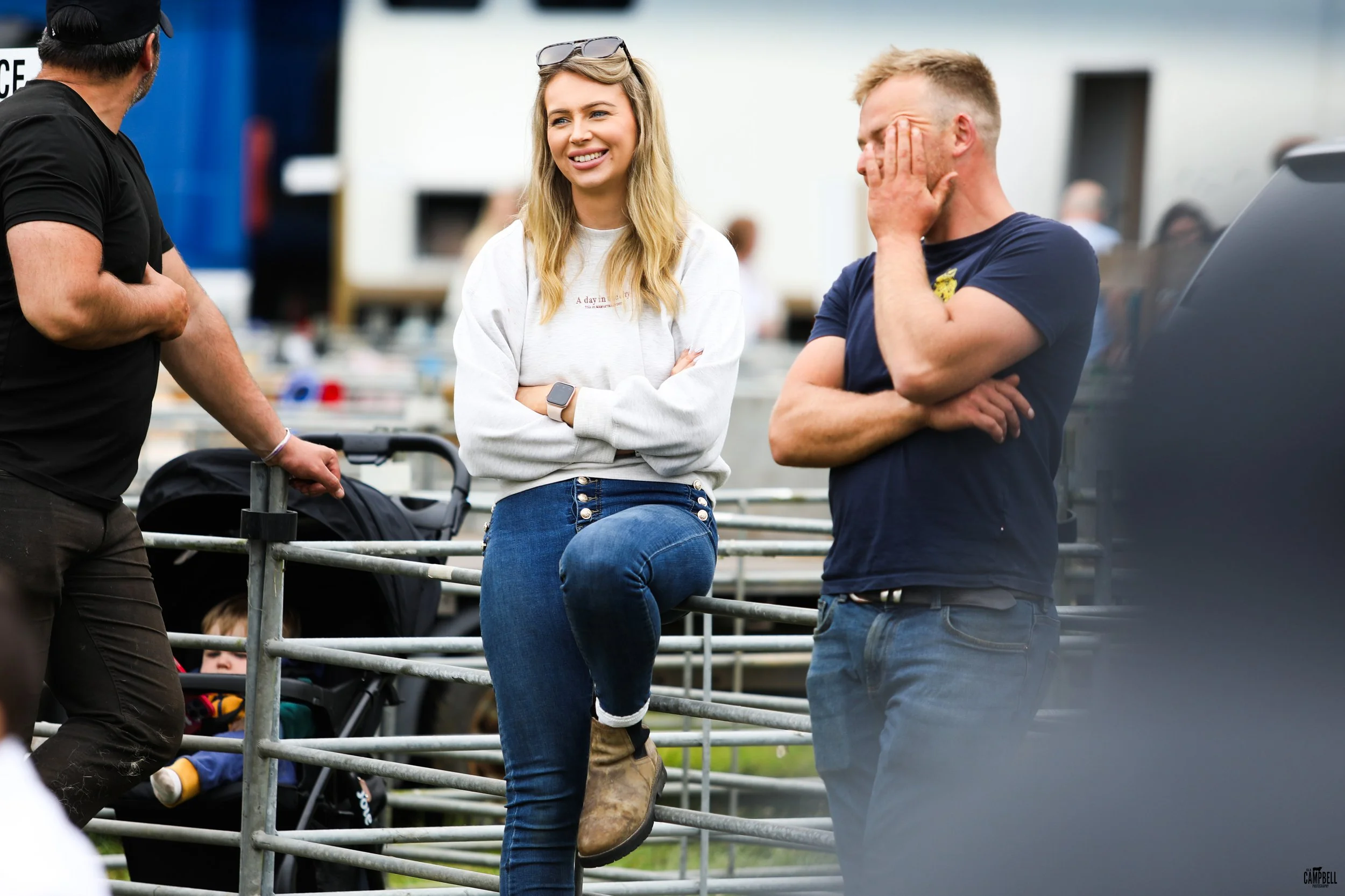 A group of people engaged in conversation outdoors, with a woman in the center smiling and wearing jeans and a white sweatshirt, surrounded by men, one of whom is covering his face while the other is turned away from the camera. There is a baby strol