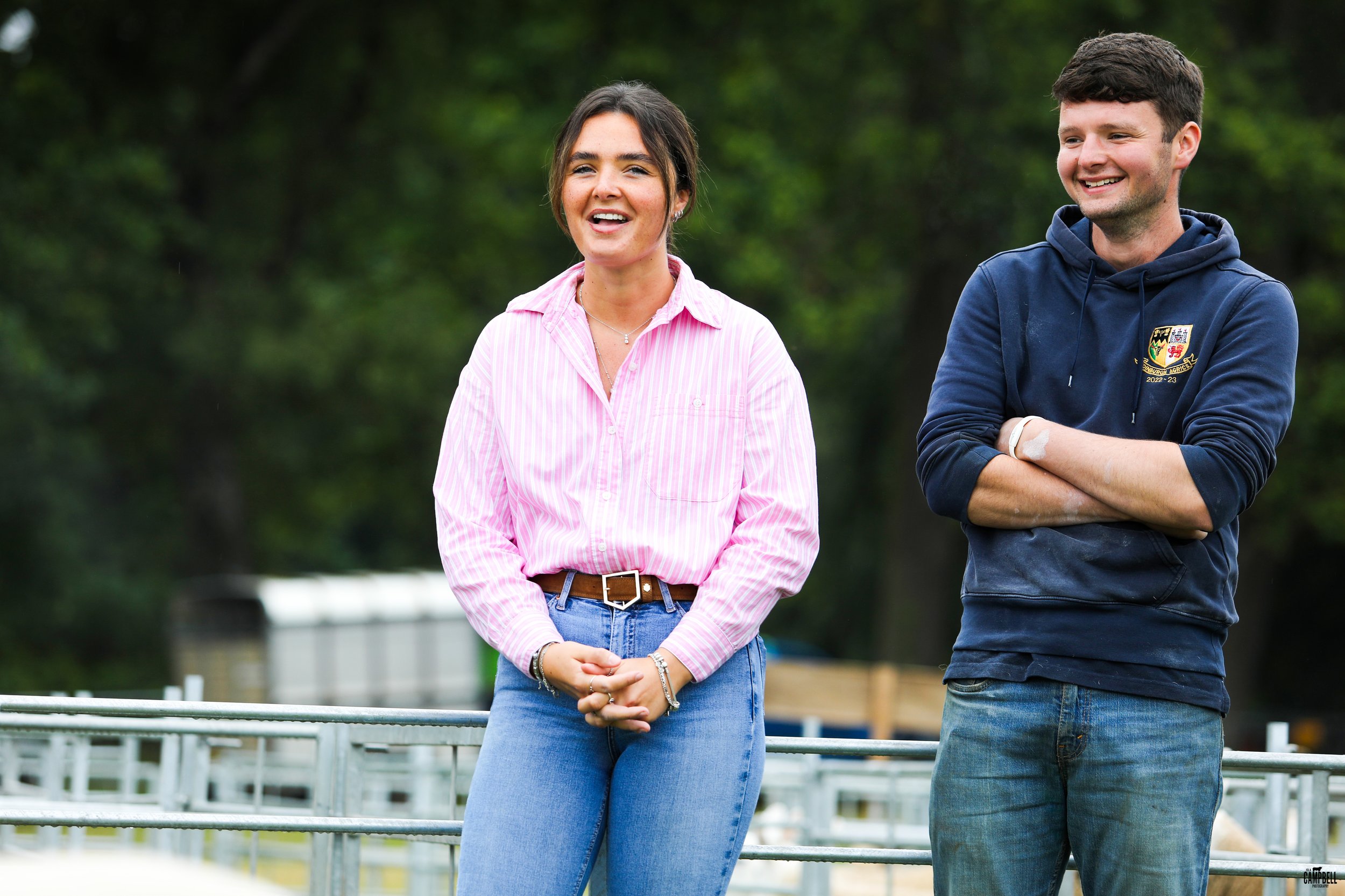 A woman wearing a pink striped shirt and blue jeans standing outdoors next to a man in a navy hoodie, smiling with his arms crossed, with a background of green trees.