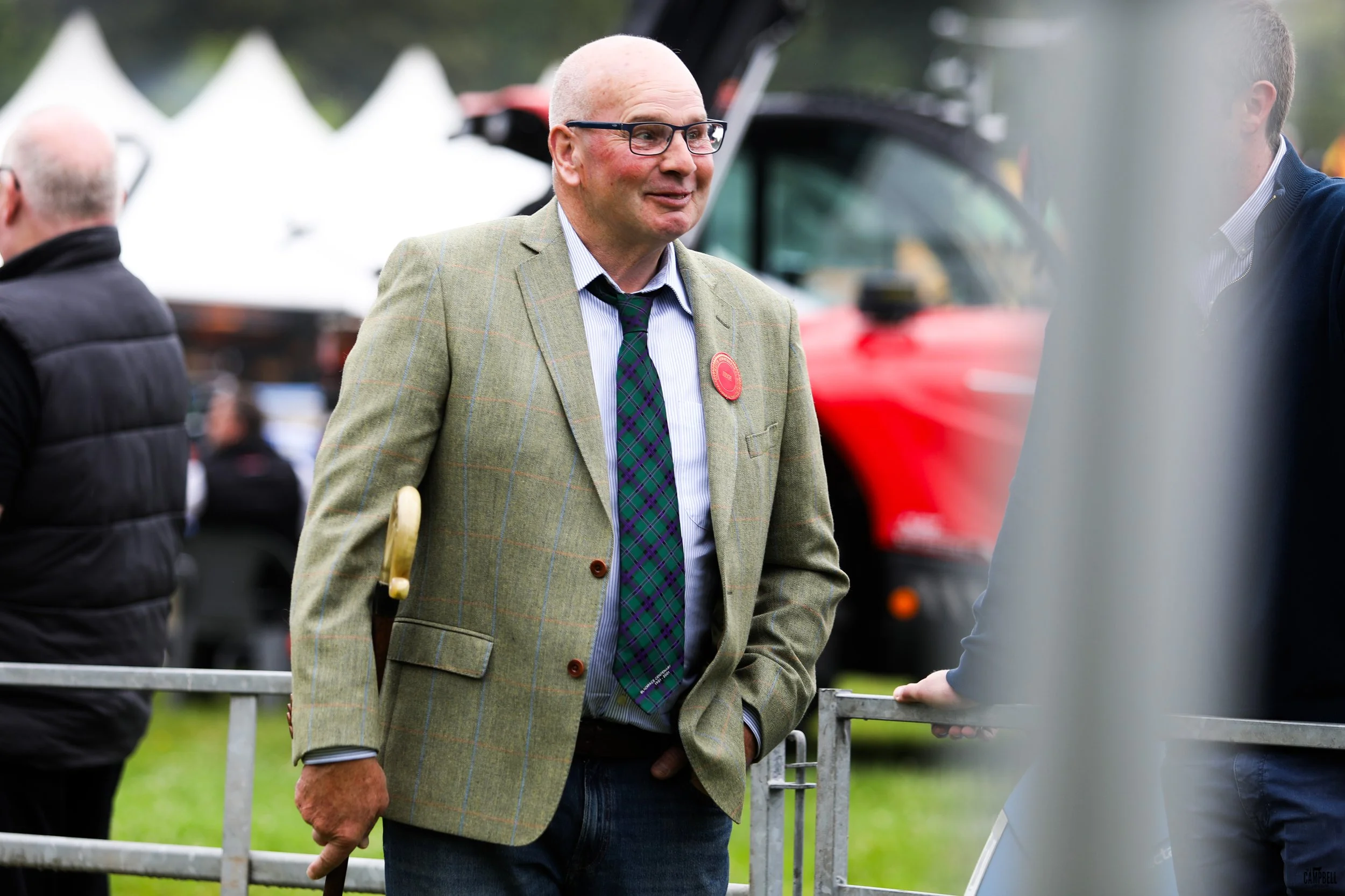 A man with glasses, wearing a green plaid blazer, white shirt, and green plaid tie, stands outdoors near a metal fence, engaging in a conversation with a person whose hand is visible on the fence. In the background, there are tents and a red vehicle,