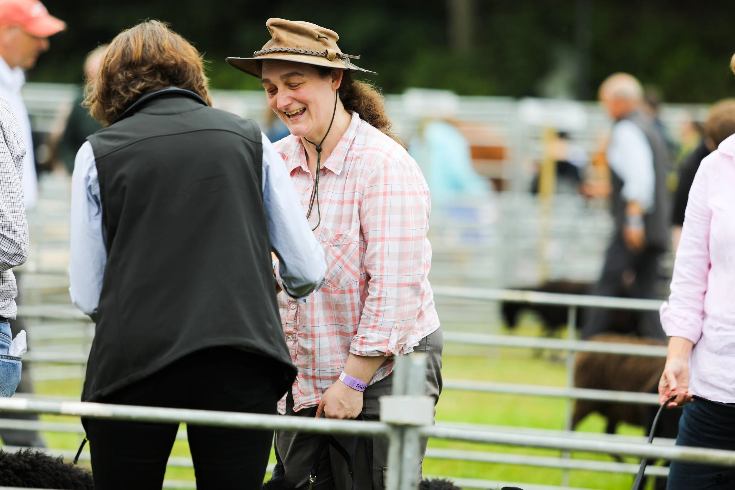 People at an outdoor event, with a woman wearing a wide-brimmed hat and a plaid shirt smiling and talking to another person wearing a black vest.