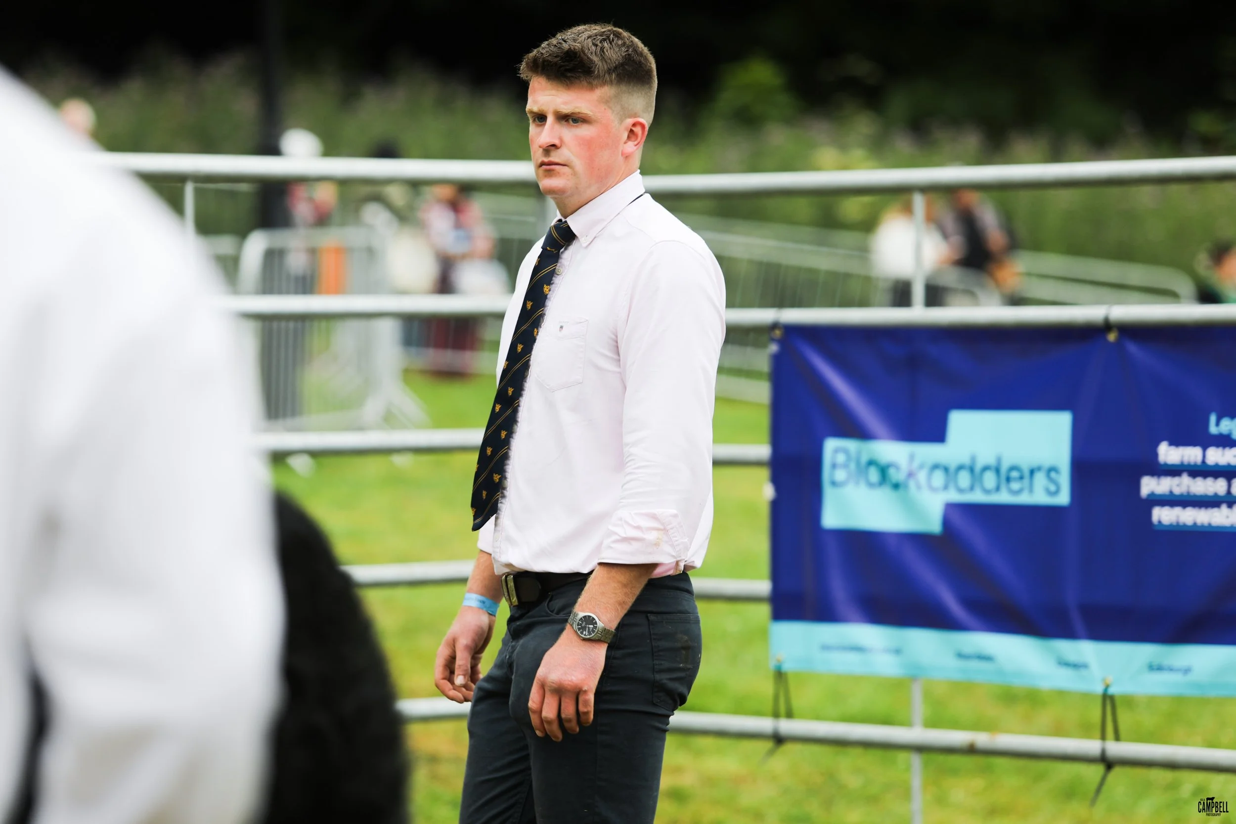 A young man with short brown hair wearing a white button-up shirt, a navy tie with small yellow designs, black pants, and a watch on his left wrist, standing outdoors with a serious expression.