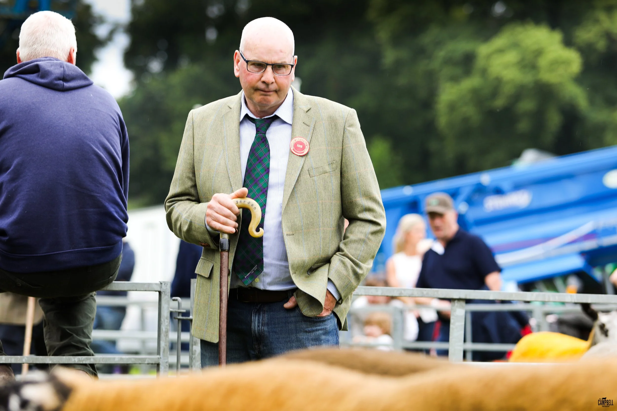 A man with glasses in a beige blazer, striped shirt, and plaid tie holding a crook, standing outdoors at a fair or market, with other people and tents in the background.