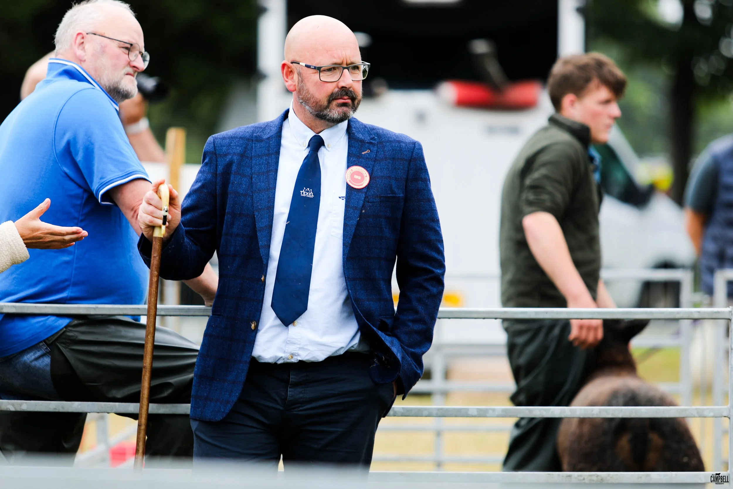 A man in a blue checkered blazer, white shirt, and black tie with a lapel pin, wearing glasses, stands with his hands in his pockets at an outdoor event, surrounded by other people.