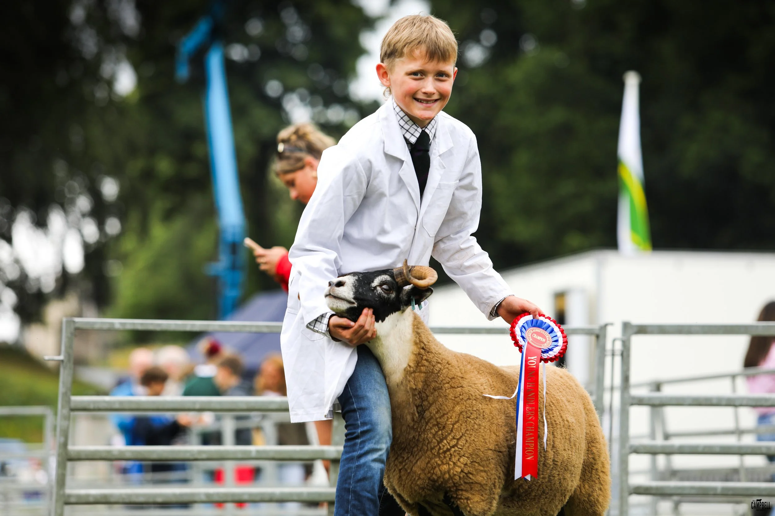 A young boy wearing a white coat and black tie is smiling while holding a ribbon and a goat at an outdoor event. There are blurred spectators and tents in the background.