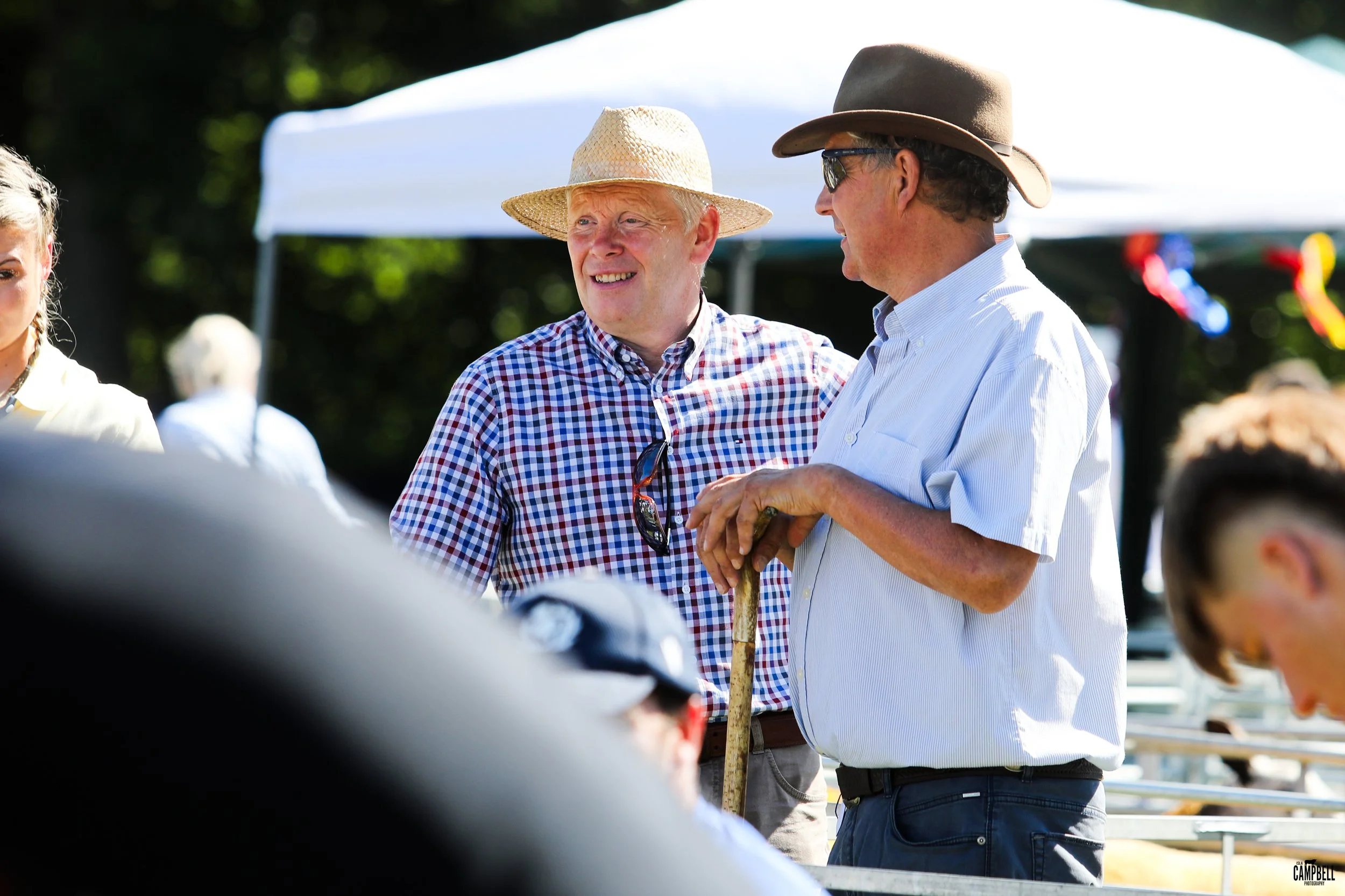 Two middle-aged men are outdoors, talking and smiling, wearing wide-brimmed hats and casual shirts. One man has a checkered shirt and glasses hanging from his collar; the other has a white shirt and a cane. There is a white tent and trees in the back