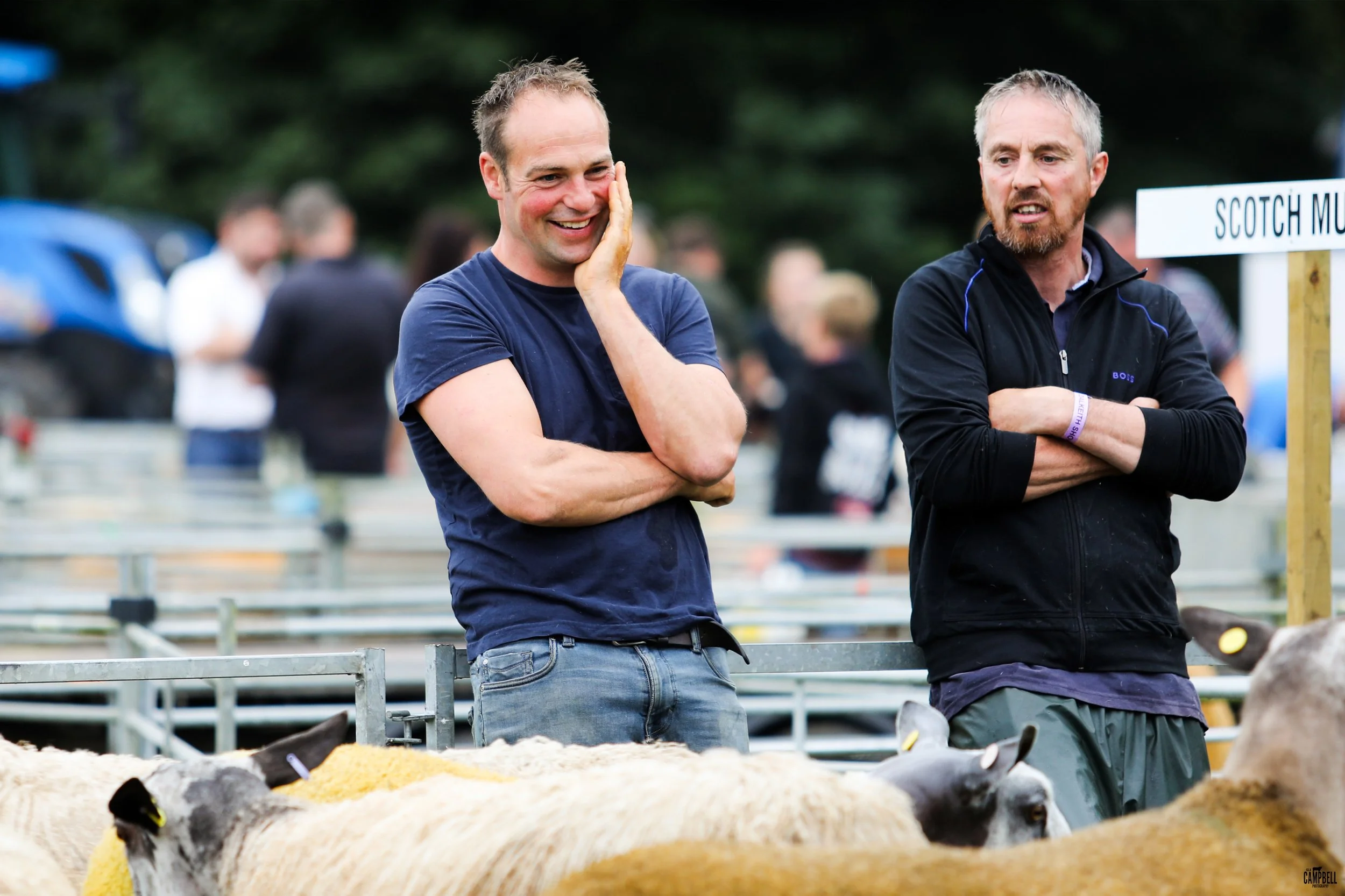 Two smiling men standing near sheep at an outdoor event, with a sign labeled 'Scotch Mutch' in the background.
