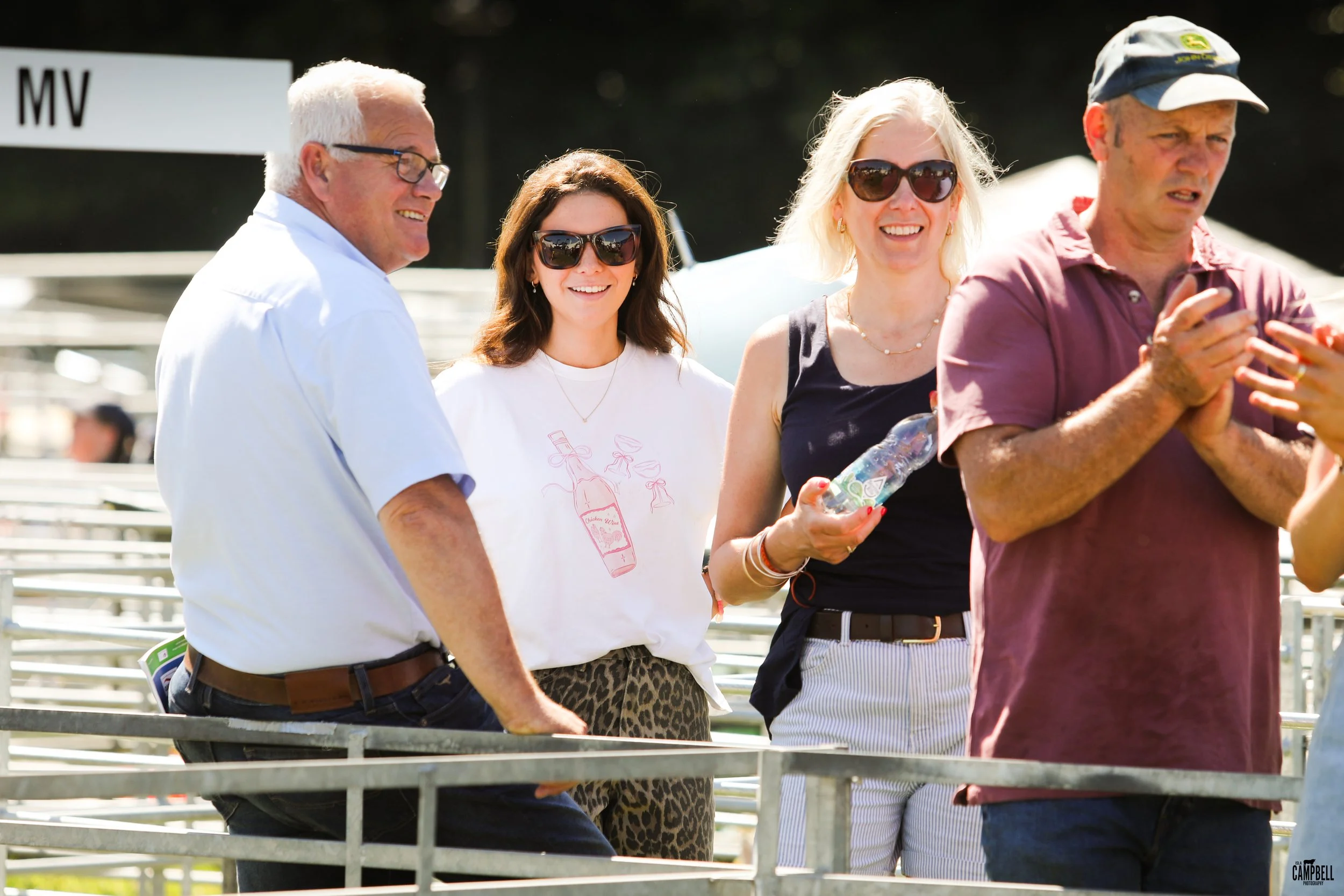 Four people, two women and two men, are outdoors and smiling. The woman on the right holds a plastic water bottle. All wear sunglasses and casual summer clothing.