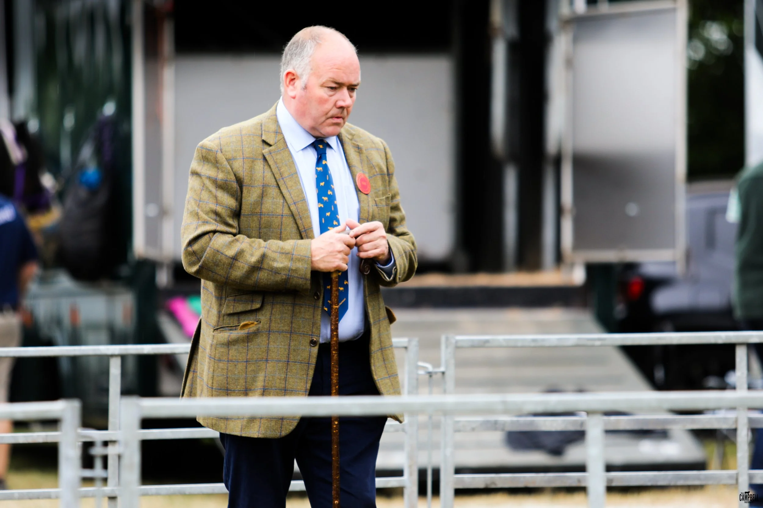 A man wearing a checked blazer, white shirt, and a blue tie with yellow patterns stands with hands clasped, looking down with a somber expression in an outdoor setting.