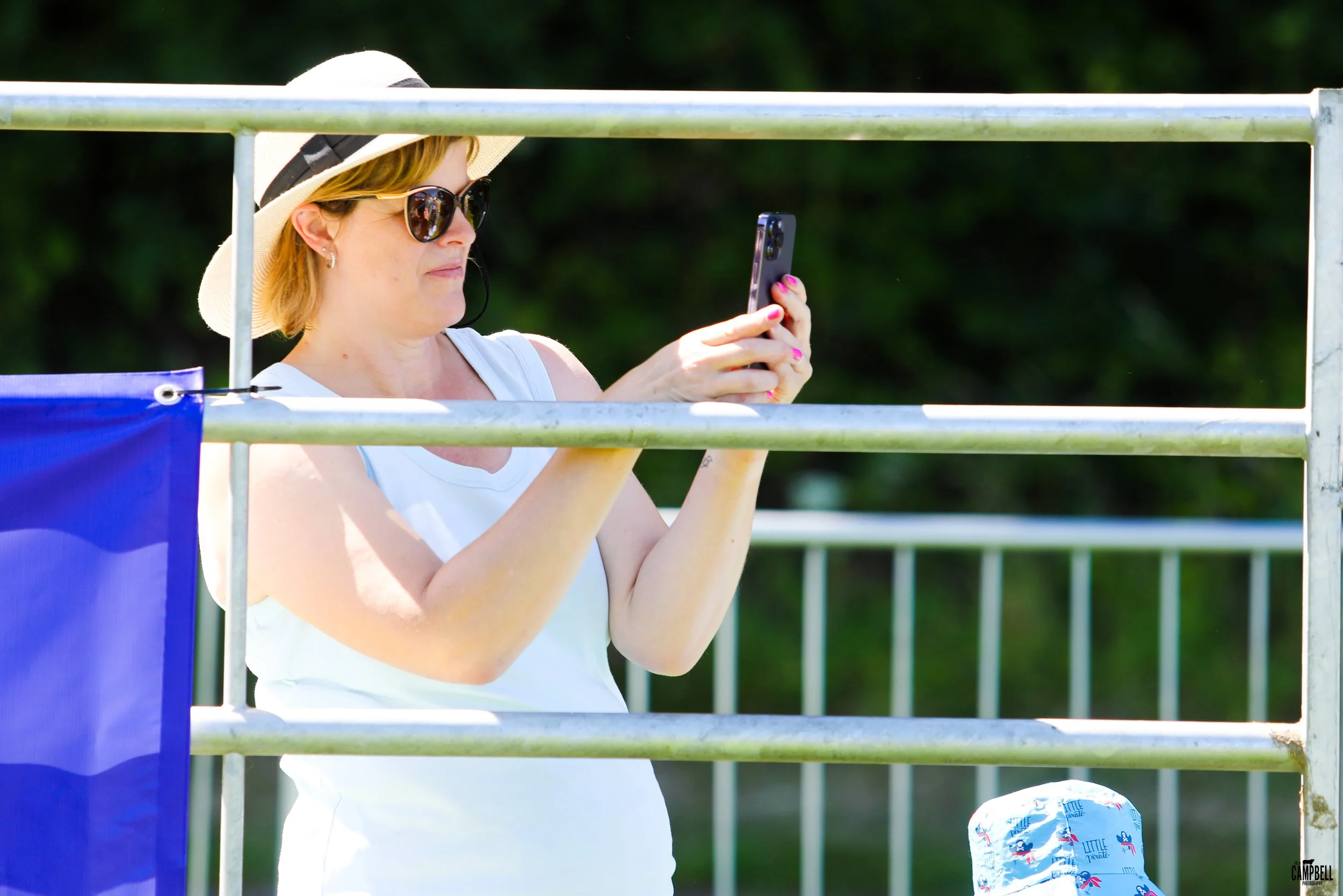 A woman wearing a wide-brim hat, sunglasses, and a sleeveless white top taking a photo with her phone at an outdoor location with metal railings and a green background.