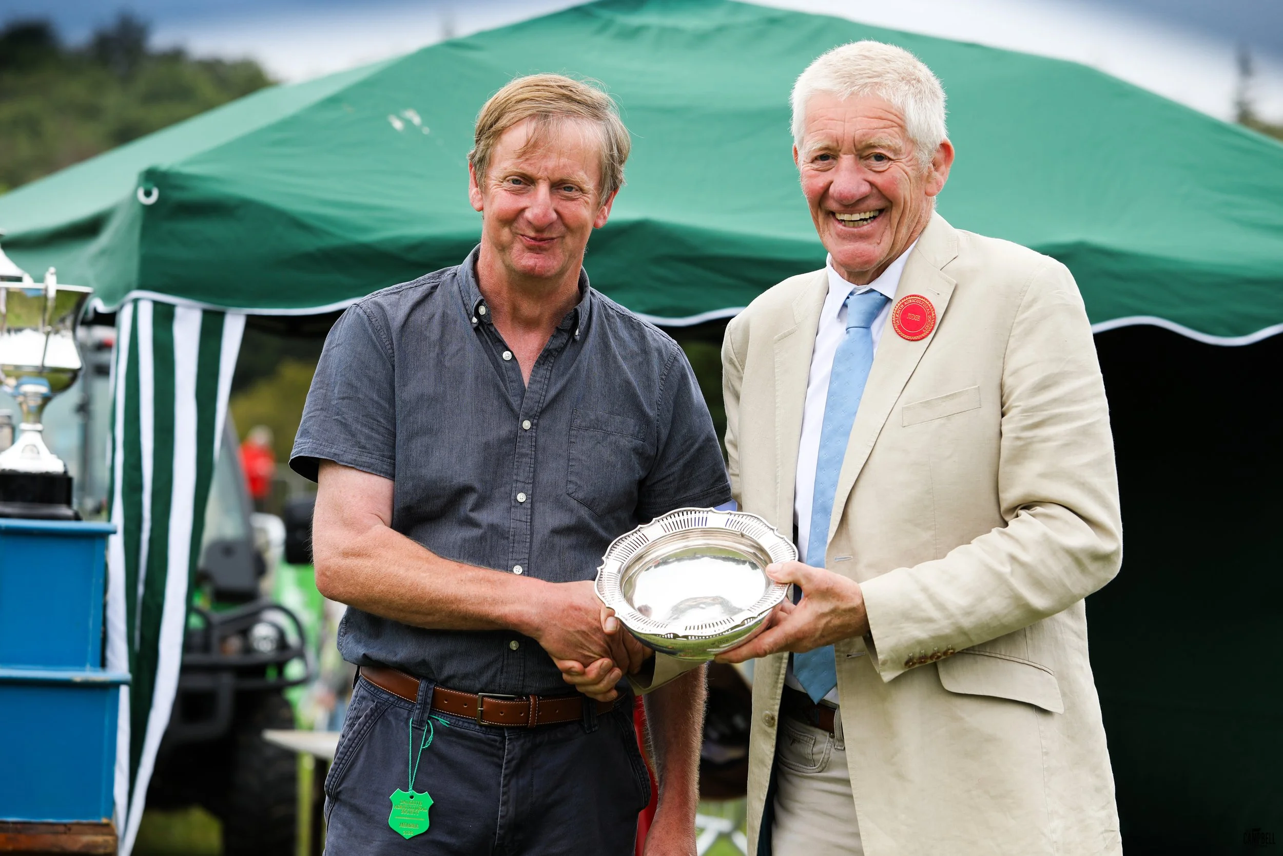 Two men smiling, one in a dark shirt and the other in a light-colored suit, are holding a silver plate together outdoors at a daytime event, with a green tent and trees in the background.