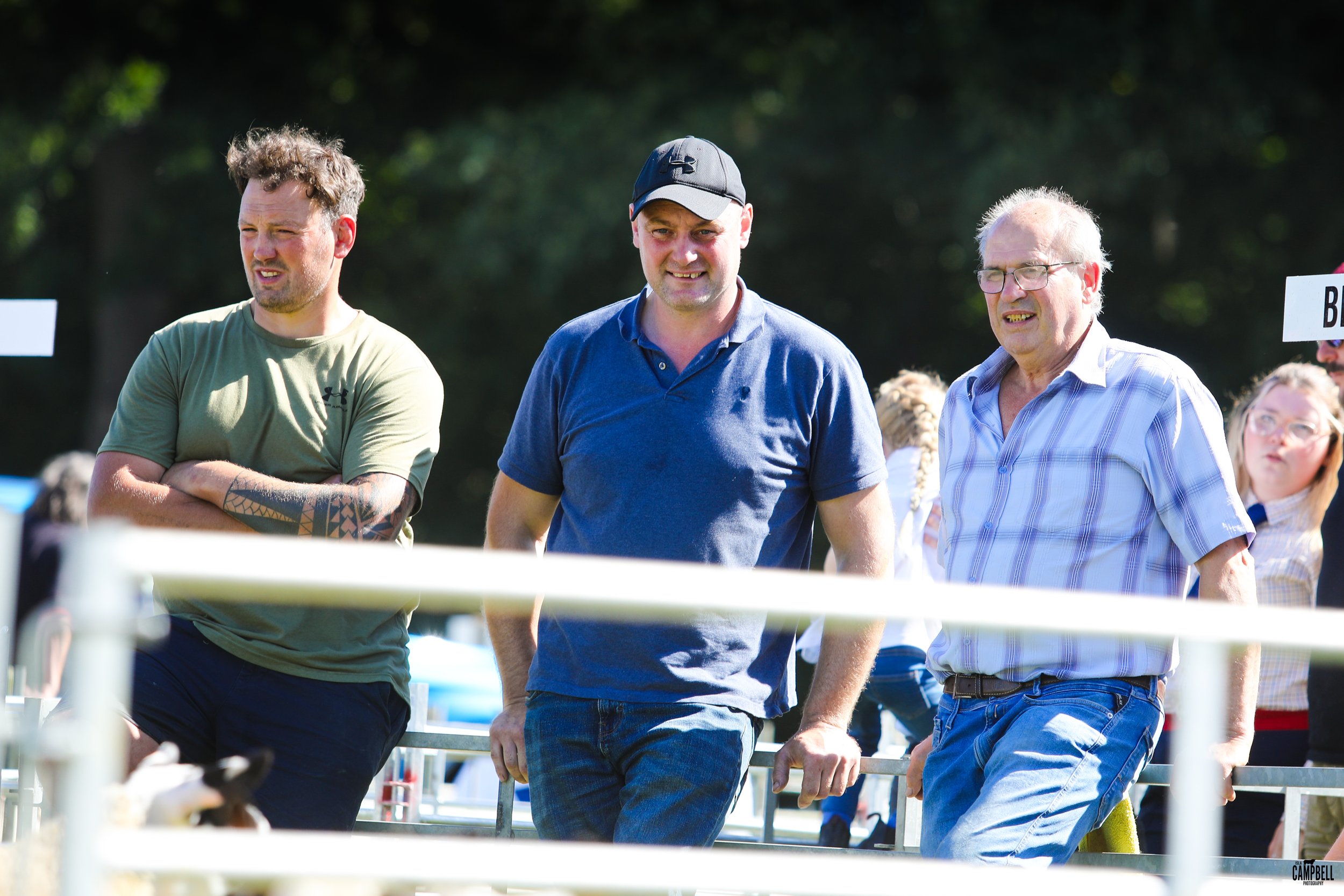 Three men standing outdoors at a daytime event, with a background of trees and other people. One man is in a green shirt with tattoos and crossed arms, another in a blue polo shirt and cap, and the third in a striped shirt and glasses, all looking at