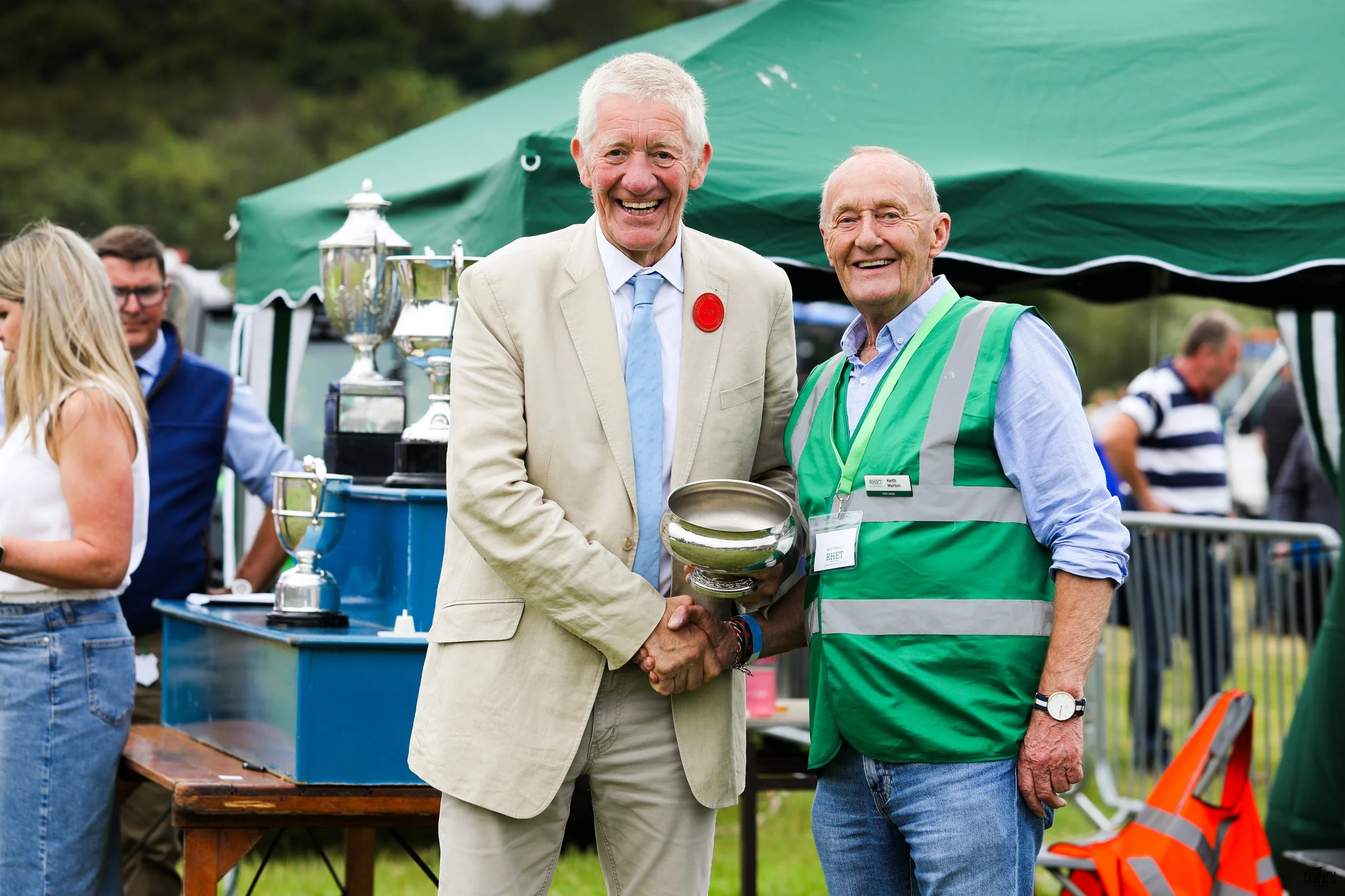 Two older men shaking hands at an outdoor event, smiling, with trophies on a table behind them. One man is in a beige suit and the other in a green vest with a name tag.